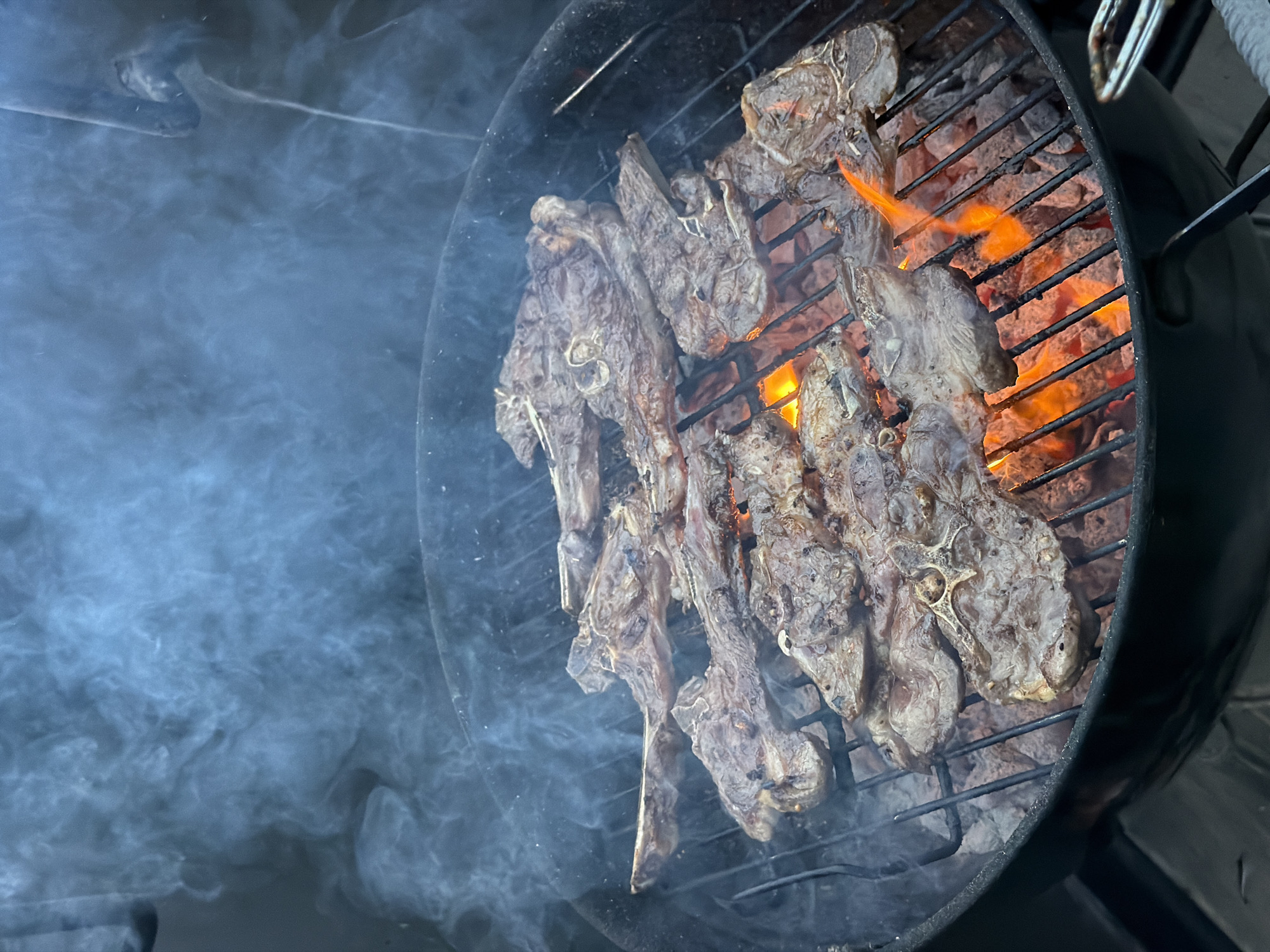 The image shows a barbecue grill with several lamb chops being cooked over an open flame. Smoke is billowing around the meat. The grill is black and round. The lamb chops are a dark brown color. The location is not specified but is purported to be in Tel-Aviv, Israel.