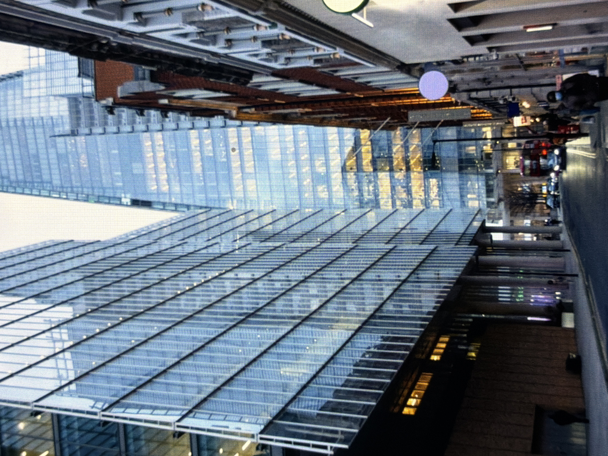 The image captures an urban street scene in London, United Kingdom, under dim lighting conditions. On the left, a tall, modern building with a facade of blue-tinted glass panels reflects ambient light. In the background, the distinctive, pointed silhouette of The Shard skyscraper is prominent, its numerous interior lights glowing golden through its glass exterior. To the right, an older, multi-story building with brick and stone elements features traditional architectural details and commercial signs, including a circular purple sign and a partial green 'A' logo. The street in the foreground features asphalt pavement with painted lines, and several vehicles, including a red double-decker bus, are visible further down the road. Pedestrians walk on both sidewalks; one individual in the foreground on the right side is seen wearing a dark jacket and a face mask. Artificial streetlights and interior building lights illuminate the scene.