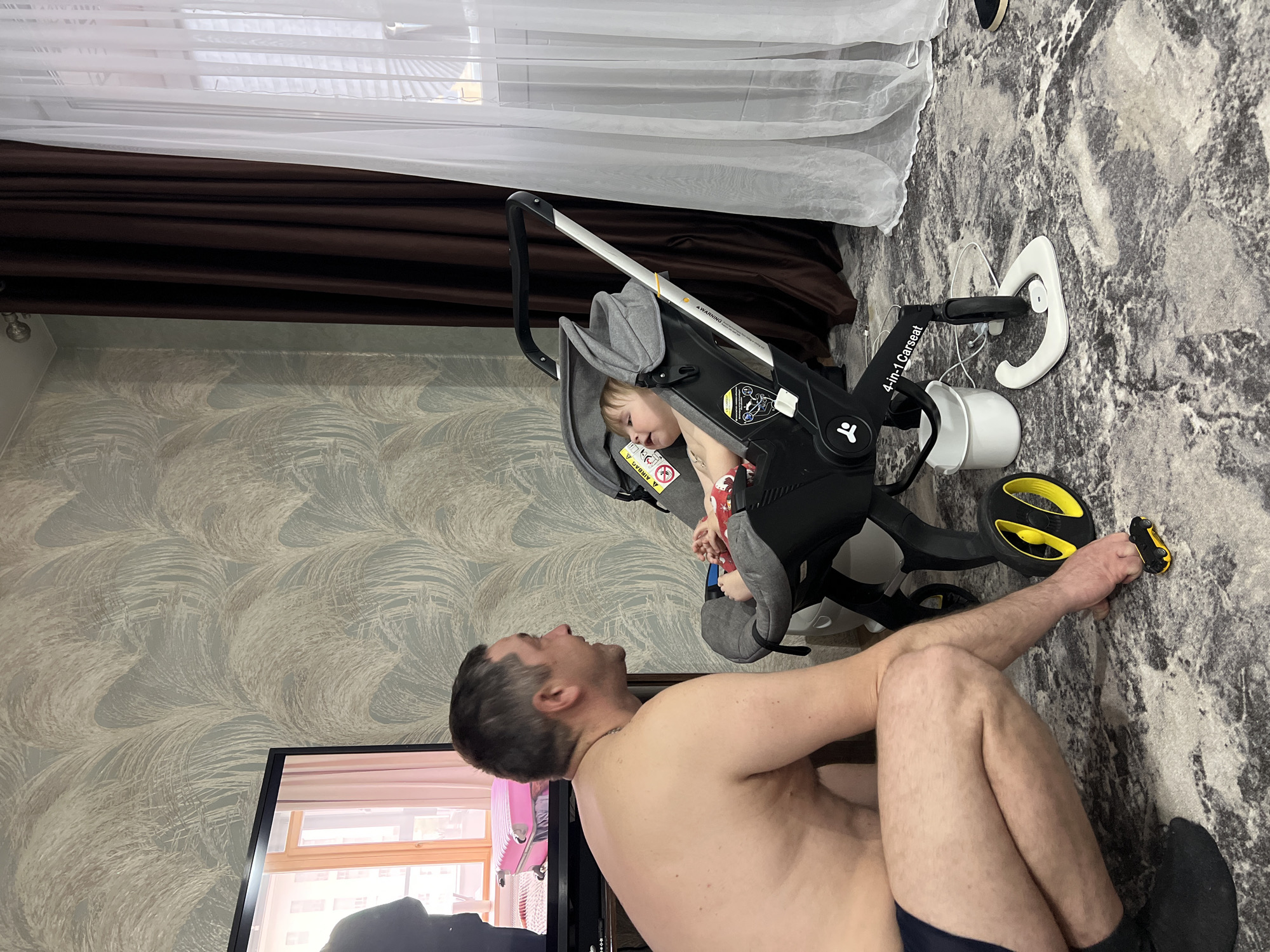 The image shows an indoor scene, possibly inside a residence in Noviy, Russia, judging by potential clues in the television screen, although there is no definitive proof of the location. A partially shirtless man is kneeling on a patterned grey and white rug. He seems to be interacting with a young child seated in a black and grey 4-in-1 car seat tricycle. The child is holding a small, possibly red, object in their hands. The tricycle is positioned near the man, suggesting an interaction. The environment includes a television set in the foreground, a patterned wallpaper behind the man, and a window with white and brown curtains in the background. The floor has a carpet with a grey and white pattern. Next to the tricycle and the man's leg, there is a small toy car.