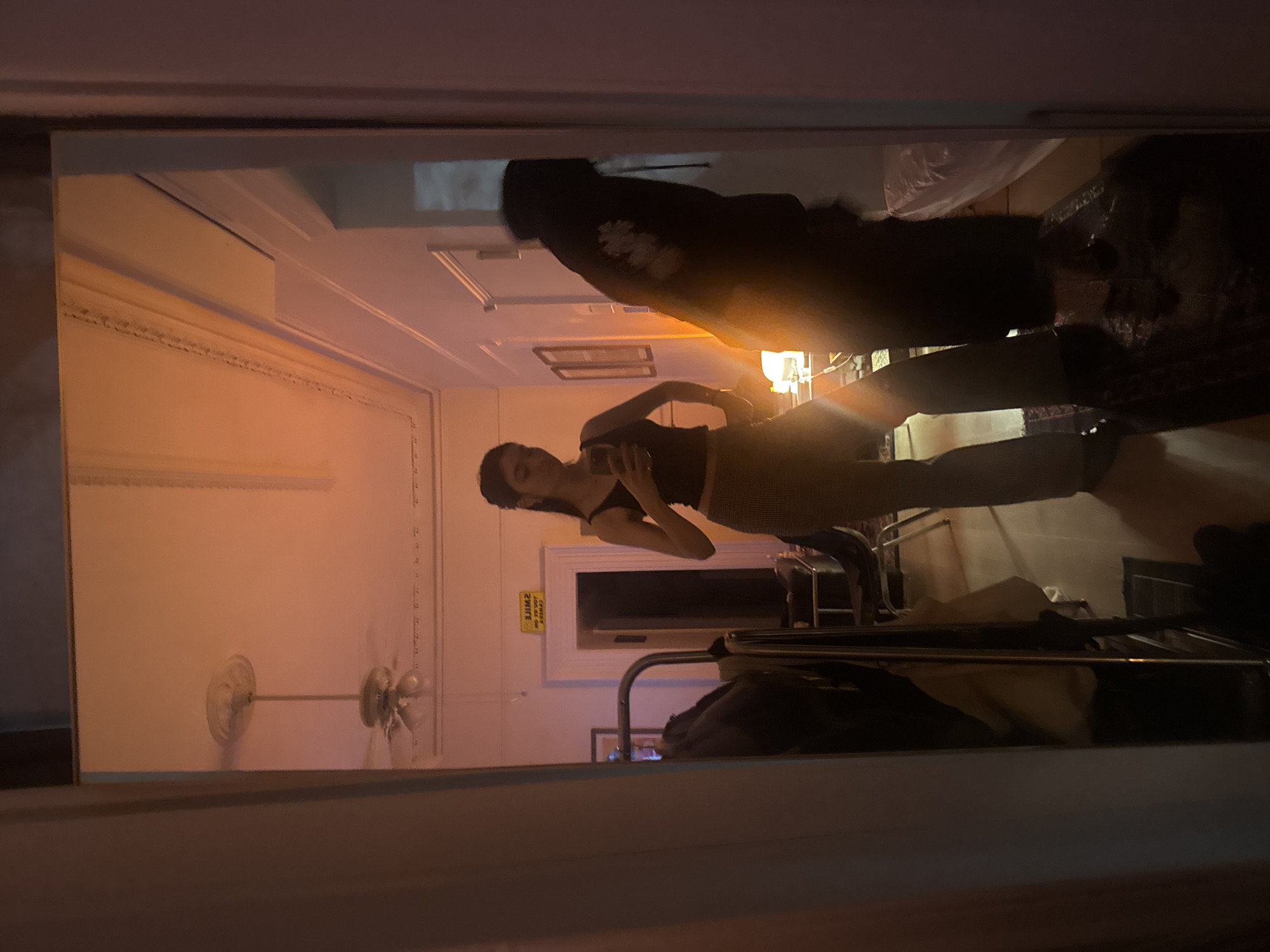 A person is taking a photo of themself in a mirror within what appears to be an apartment. The apartment features a ceiling fan, ornate molding, and various pieces of furniture. A second person is present in the mirror's reflection, further back in the room. Without further context, a specific location in New York City cannot be determined.