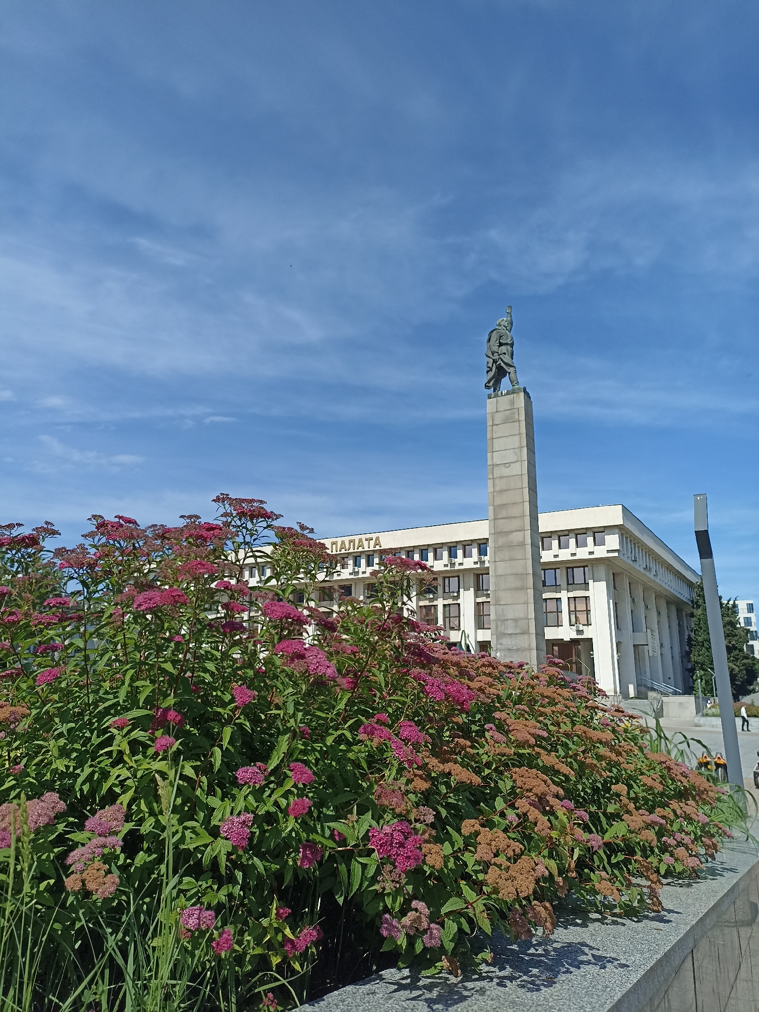 In Burgas, Bulgaria, a statue of a soldier stands tall on a pedestal in front of a modern, light-colored building. The building has a sign with Cyrillic lettering that reads "ПАЛАТА," indicating it is a palace or official building. In the foreground, a lush bush of pink and brown flowers creates a vibrant border. The sky is a clear blue with scattered clouds. Two people can be seen in the background, suggesting this is a public space.