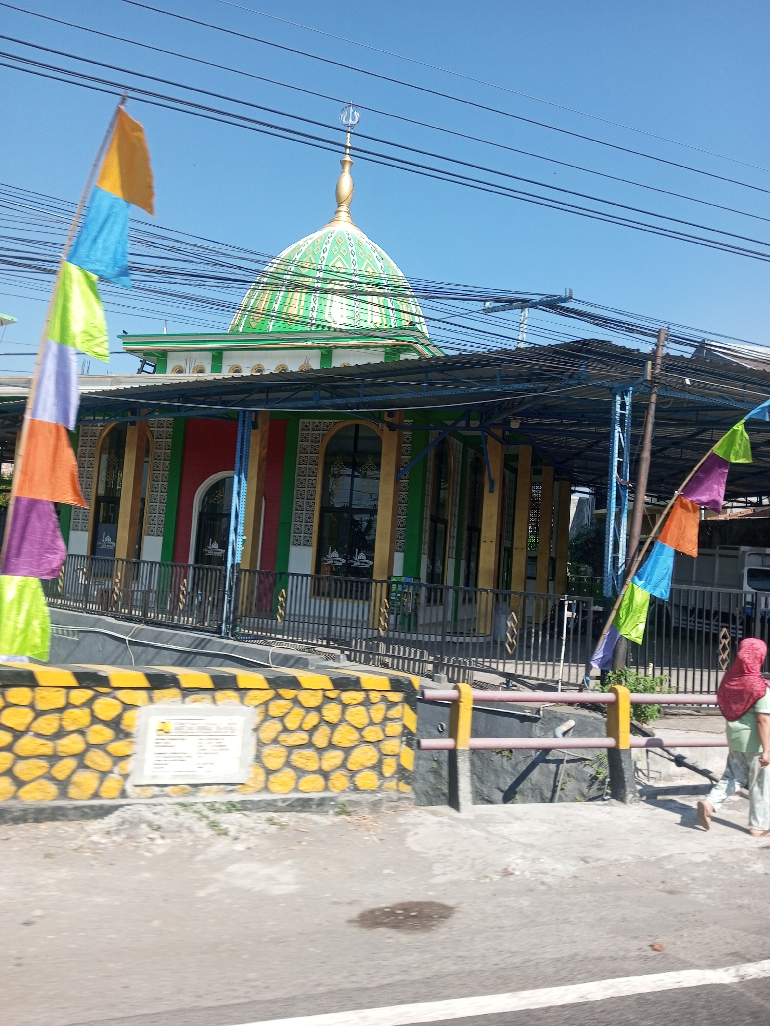 In the city of Sengon Agung, Indonesia, a mosque with a distinctive green dome topped with a crescent moon stands under a clear blue sky. The mosque features colorful decorations, including red and green accents on its facade, and is partially shaded by a blue-roofed canopy. Colorful flags flutter in the foreground, adding a vibrant touch to the scene. A yellow and black striped wall with a stone pattern separates the mosque area from the road. A sign is mounted on this wall. A person wearing a red headscarf and light-colored clothing walks away from the camera on a concrete walkway adjacent to the road. The environment appears to be a street scene with power lines overhead and a dry, paved surface.