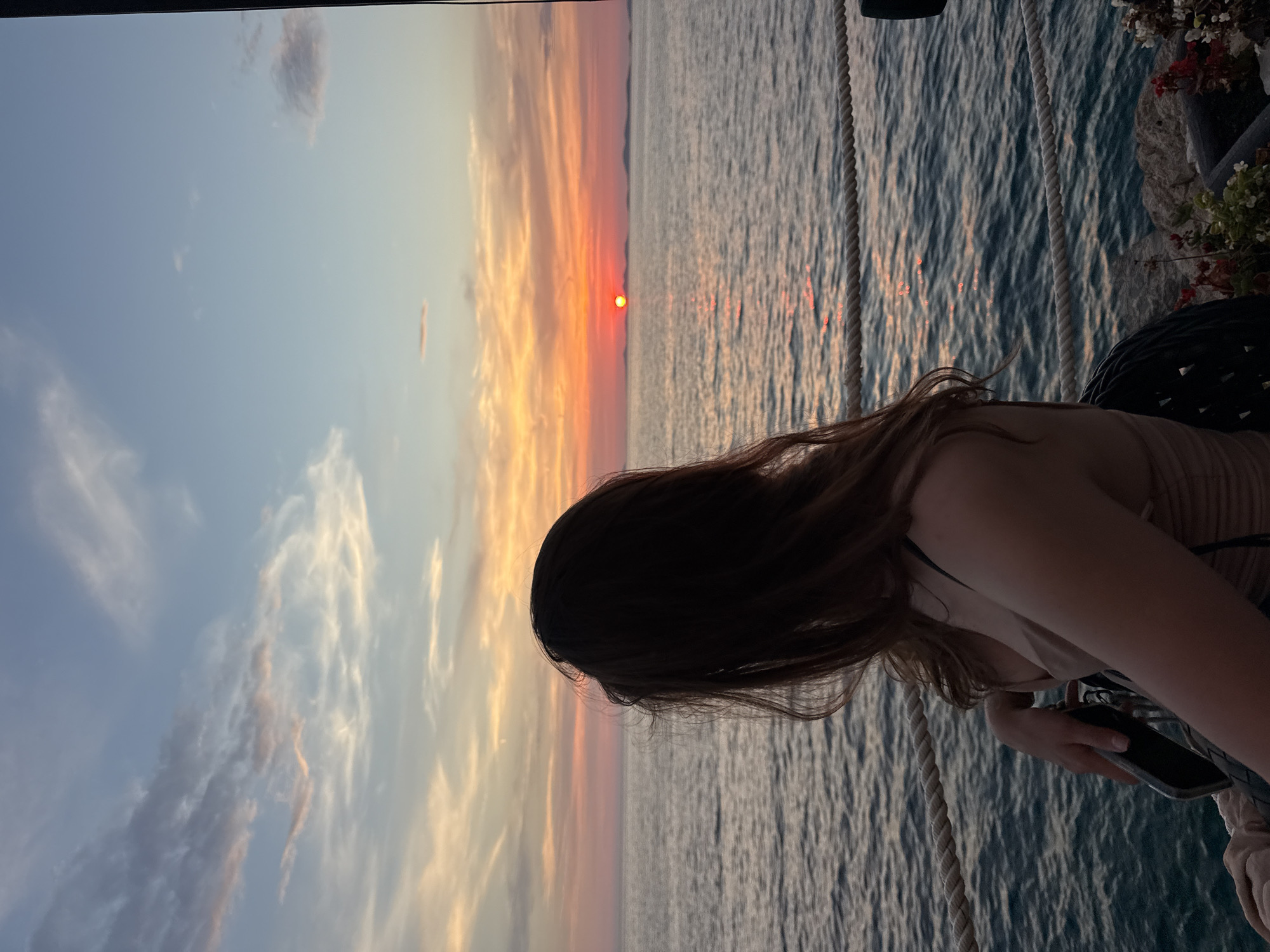 The image captures a young woman with long, dark brown hair, viewed from behind, as she gazes out at a sunset over the Adriatic Sea in Primošten, Croatia. She is wearing a light-colored, ruched dress and is seated, with her left arm resting on a railing. The sea is a deep blue, with gentle waves reflecting the warm hues of the setting sun. In the distance, a sliver of land or islands is visible on the horizon. The sky is a gradient of colors, transitioning from a pale blue at the top to shades of orange, pink, and yellow near the horizon. A rope railing runs horizontally across the foreground, separating the seating area from the sea. The scene evokes a sense of tranquility and beauty, characteristic of a Mediterranean sunset.