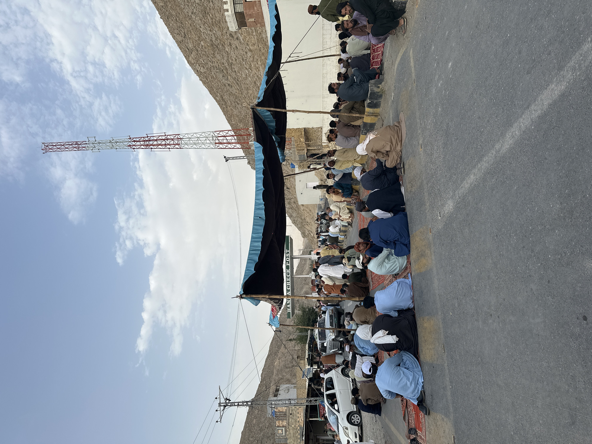 A crowd of men are gathered on a paved road in front of a check post under a canopy in Quetta City Tehsil, Pakistan. The men are sitting on rugs laid out on the road. Many are wearing traditional Pakistani clothing such as shalwar kameez. Several vehicles are parked behind the check post, including a white car and a silver SUV. A red and white cell tower stands tall in the background, with a rocky hillside to the right. The sky is partly cloudy.
