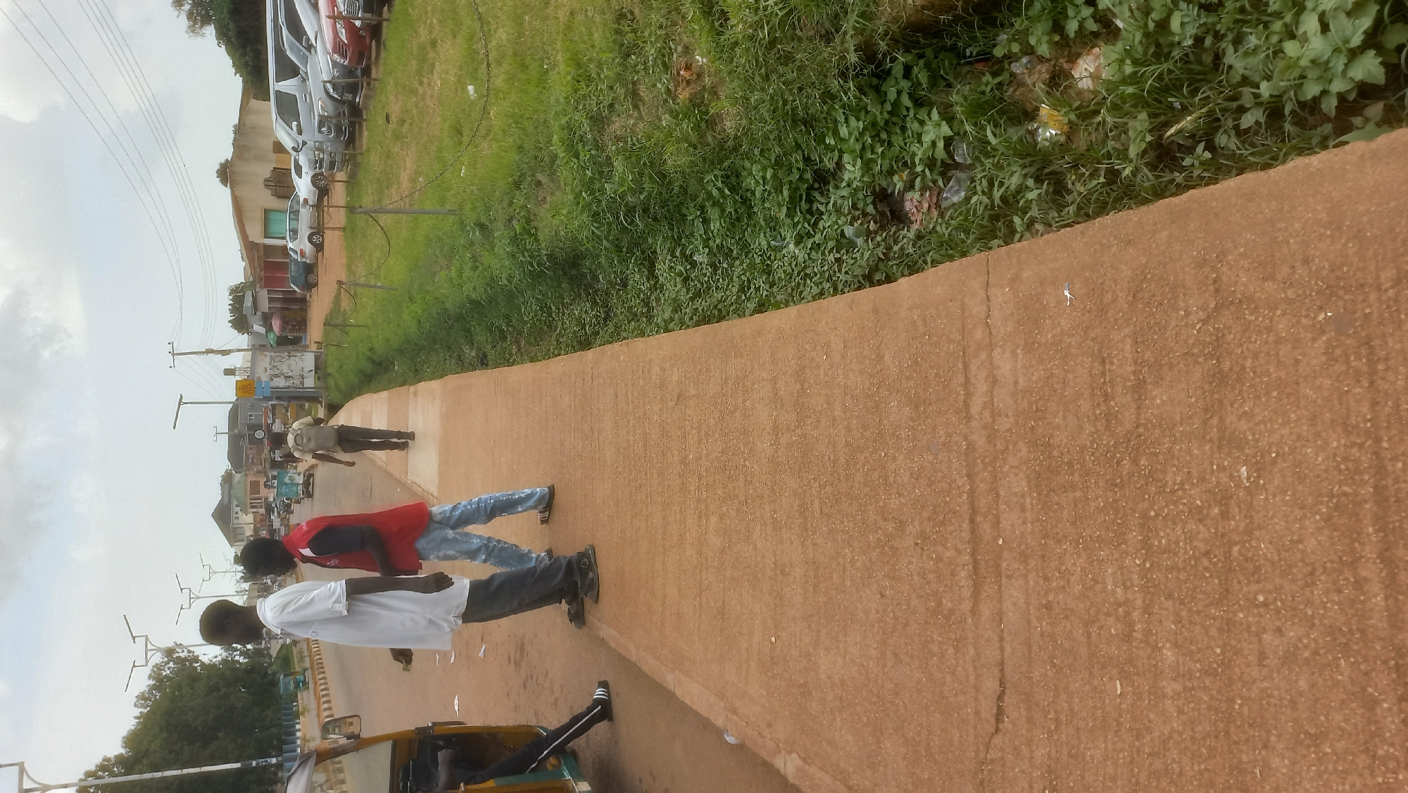 In Gura Top, Nigeria, a street scene unfolds under an overcast sky. A concrete sidewalk runs along the right side of the image, bordering a grassy area with a shallow ditch filled with vegetation and some litter. On the sidewalk, three individuals are visible. Two young men stand near a bright yellow and green auto-rickshaw parked on the left edge of the frame, partially obscuring it. The man on the left wears a white shirt and dark pants, while the man in the middle sports a red vest over a blue shirt and blue jeans. Further down the sidewalk, a third person, a young male with a backpack, walks away from the camera. Across the street, a line of parked cars, including silver SUVs and sedans, are visible, with a building and shops lining the opposite side of the road. Streetlights and utility poles are present along the street.