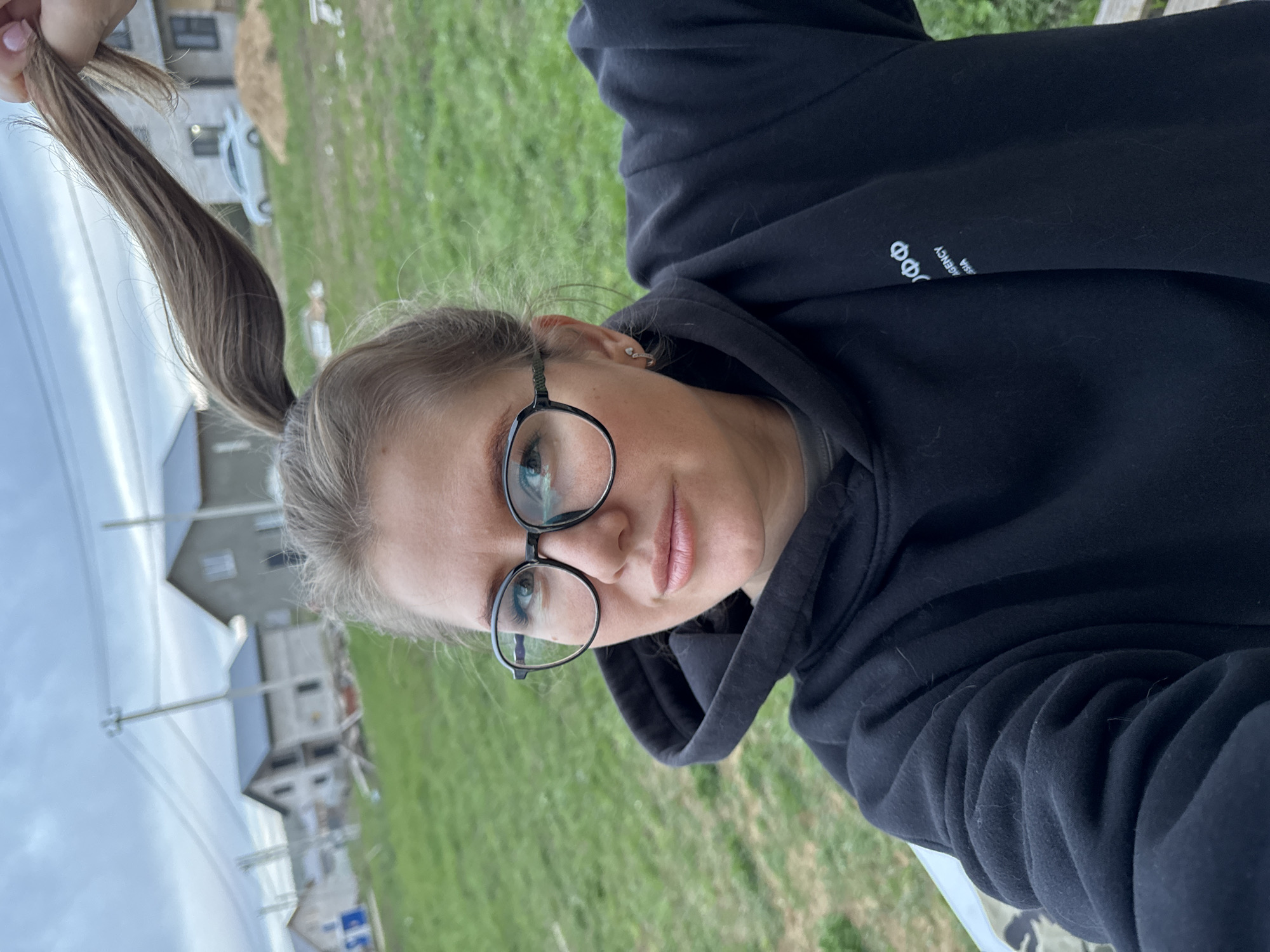 A woman with light brown hair pulled into a ponytail is shown from a slightly high-angle perspective, seemingly holding her hair up. She is wearing black-rimmed glasses and a black hooded sweatshirt with white lettering on the sleeve. The background shows a grassy field with houses under construction in the distance. The sky is overcast. A white car is partially visible near one of the houses. The location is the city of Ферма, Russia.