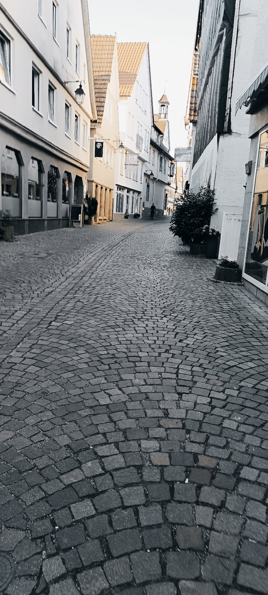 The image shows a narrow cobblestone street in Winnenden, Germany. The street is lined with buildings on both sides, featuring traditional European architecture with light-colored facades and tiled roofs. Some buildings have shop fronts at ground level, with arched windows. A single pedestrian, dressed in dark clothing, is walking away from the camera down the street. There are trees and planters placed along the buildings. The sky is overcast, and the overall lighting suggests it might be late afternoon or early morning. The foreground is dominated by the textured cobblestone pavement.