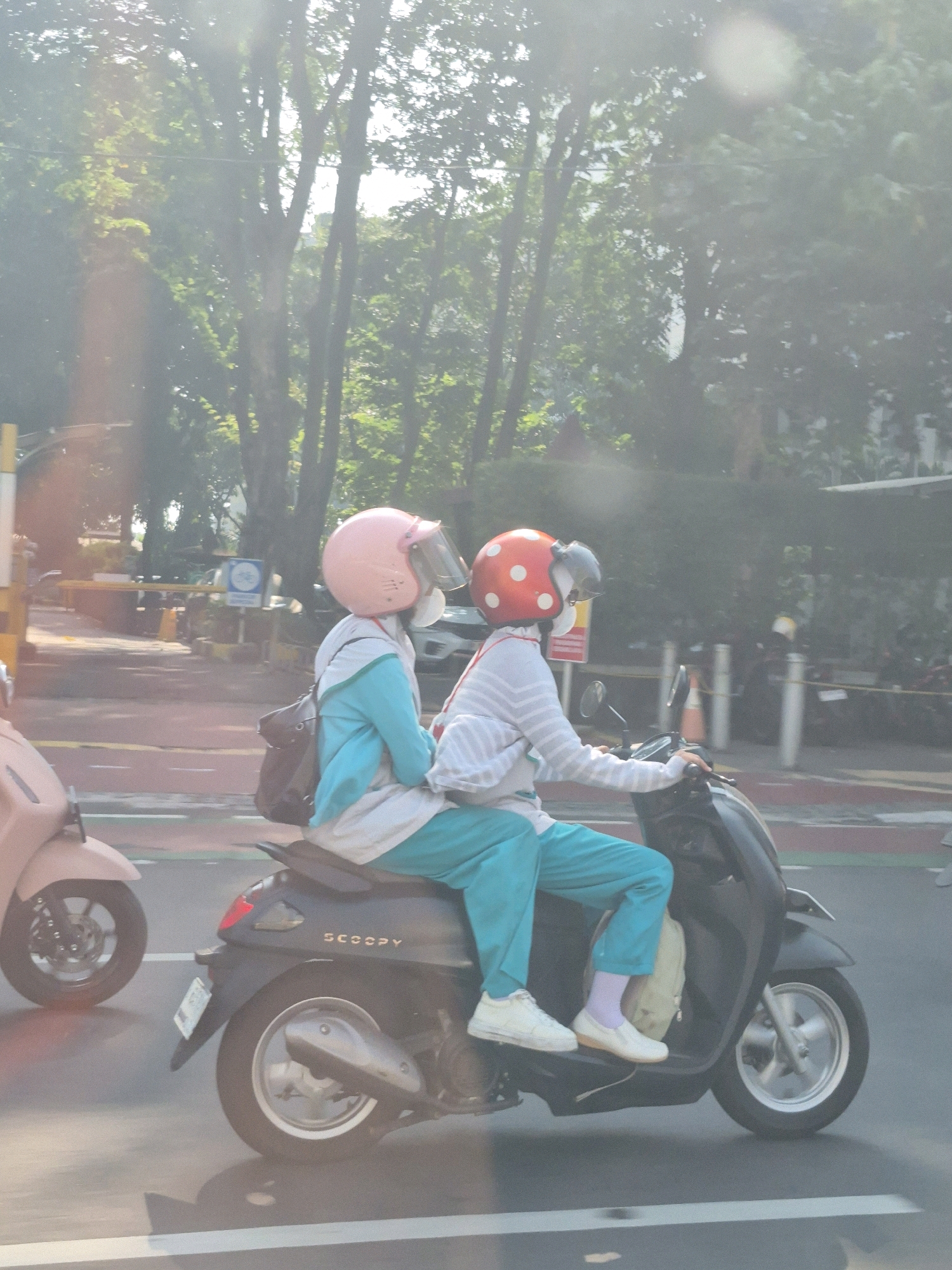 Two individuals are riding a black Honda Scoopy motorcycle on a city street. The person in the front is wearing a pink helmet and a teal and white outfit. They are carrying a dark backpack. The person in the back is wearing a red helmet with white polka dots and a striped long-sleeved shirt with teal pants. Both individuals appear to be wearing masks. A light pink scooter is visible to the left of the frame, also on the street. The background consists of trees and foliage, suggesting a location with greenery within the city. The lighting indicates it is daytime, possibly morning or afternoon due to the dappled sunlight filtering through the leaves. The road surface appears to be asphalt with white lane markings. A yellow barrier and a sign with a bicycle symbol are visible in the background on the left side of the street.