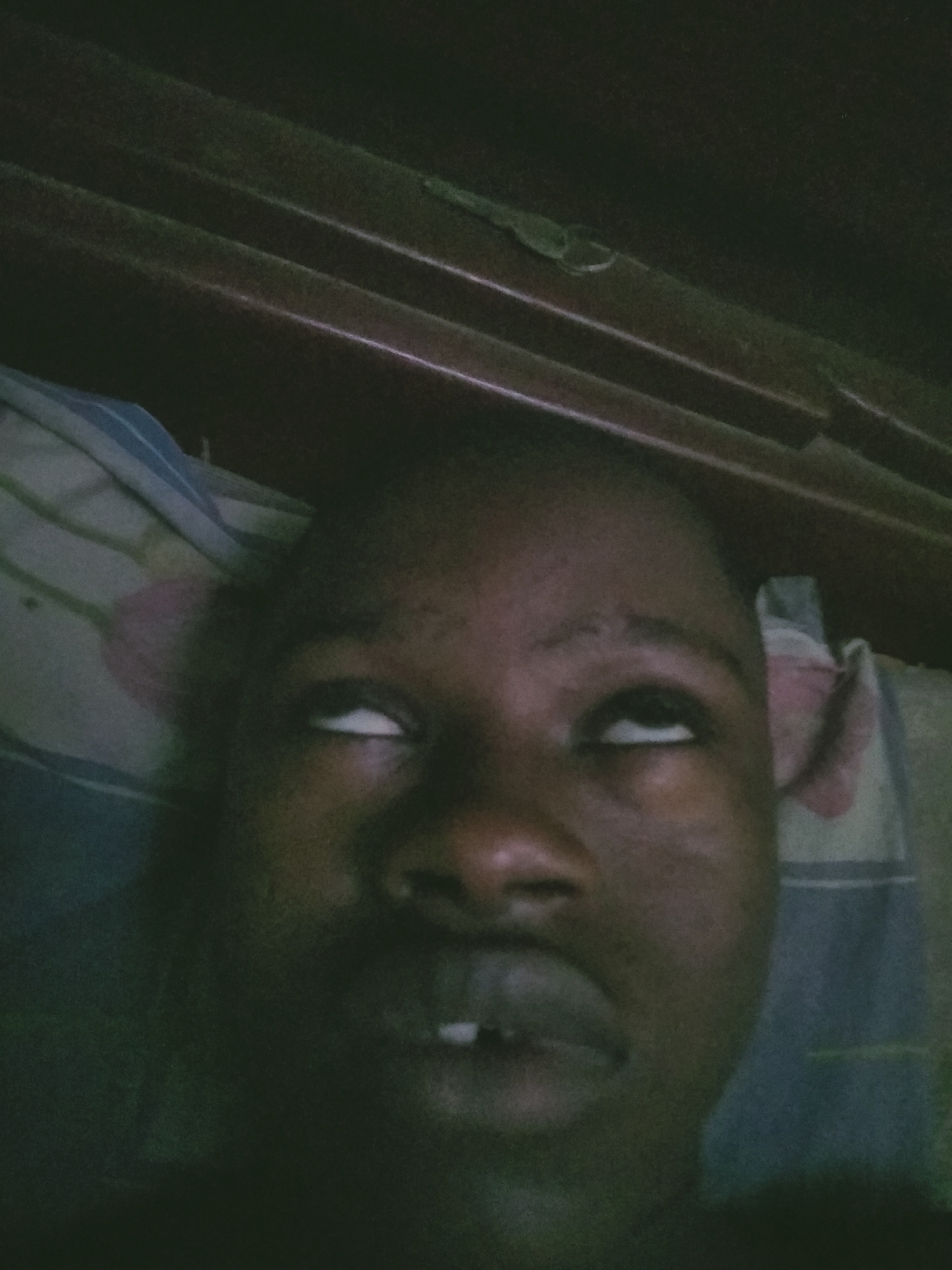 A low-light, close-up portrait shows a person lying down and looking upwards. The individual has dark skin and is captured with their eyes rolled back, exposing only the whites, while their mouth is slightly open, revealing a gap between their two front teeth. This expression appears exaggerated, possibly for comedic effect.
The person's head rests on what seems to be a pillow or bedding with a striped pattern that includes shades of blue, white, and a muted pinkish tone. Above their head, a dark wooden structure with horizontal ridges or slats is visible, suggesting a bed frame, headboard, or the underside of a bunk bed. A small, metallic hook or clip can be faintly seen attached to this wooden structure in the upper left area of the image. The lighting is dim and muted, casting shadows that suggest an indoor setting, possibly a bedroom, at night or in low-light conditions. No text is visible in the image.