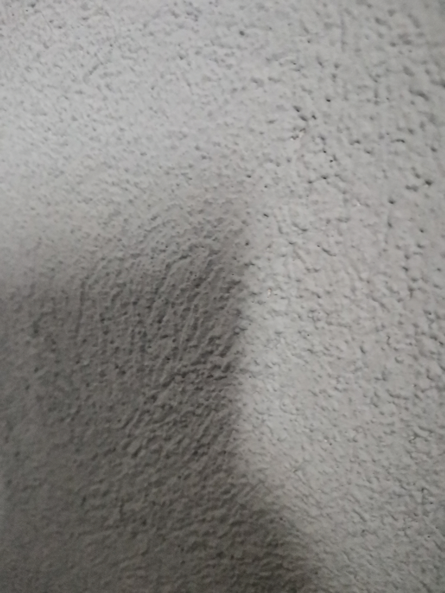 The image shows a close-up of a textured, light gray surface. The texture is irregular and rough, with small bumps and indentations. The lighting is uneven, with a darker area on the left and a lighter area on the right. No specific details indicate the location as Pokrovnik, Bulgaria. The image contains no people, subjects, objects, or actions beyond the textural surface itself. There are no observable interactions.