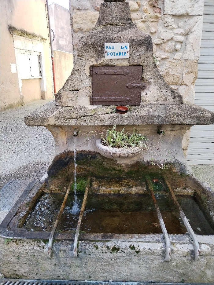 An old stone public drinking fountain, labeled "Eau Potable" (Drinking Water) in French, features a metal door above a decorative plant in a pot. Water flows from a metal faucet into a mossy stone basin divided into sections by metal bars. The fountain is situated on a narrow stone street, adjacent to buildings with white siding and a closed shutter.