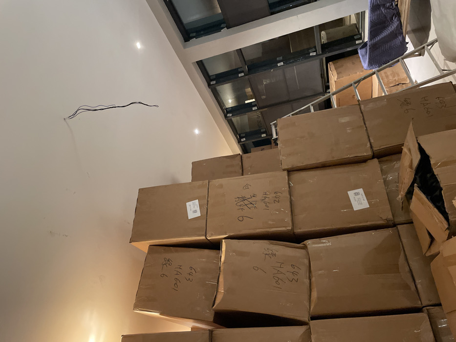 The image shows a stack of cardboard boxes reaching towards the ceiling. Many boxes are labeled with handwritten characters, including "643 HA601 绿6". A few boxes are partially open. Electrical wiring extends from the wall. Above the boxes, a series of glass panels are visible in the ceiling, along with a ladder and a draped object. The overall scene suggests a storage area or delivery location.