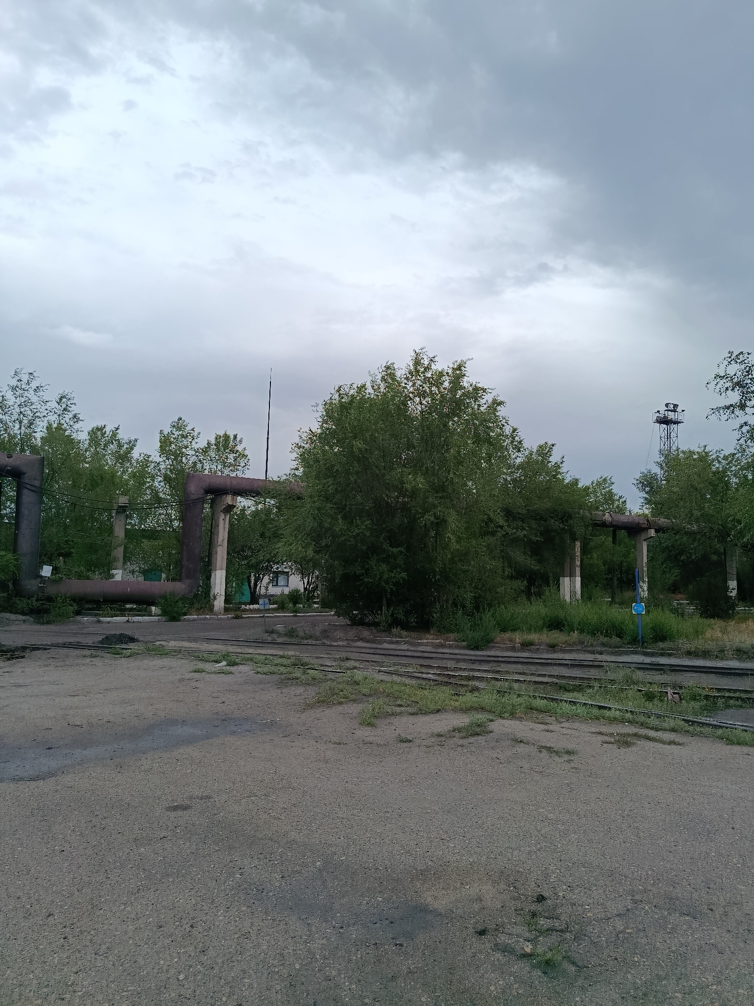 The image depicts an outdoor scene with industrial infrastructure and natural elements under a cloudy sky. In the foreground, a grey, uneven paved surface is visible, transitioning into a gravel and grass area with multiple sets of parallel railway tracks.
Dominating the midground are large, weathered industrial pipes supported by concrete pillars. These pipes appear to be part of an old or disused industrial facility. Dense green foliage, including trees and bushes, surrounds the pipes and partially obscures structures behind them. A white building with a dark-framed window is visible through an opening in the pipes.
To the right, a tall, dark metal tower with lights and structures at its top stands against the sky. A blue sign with white lettering is mounted on a post near the railway tracks. The sky is overcast with shades of grey and white clouds. The overall impression is that of an industrial area in a state of partial disrepair, integrated with overgrown vegetation.
This scene is located in the city of Saran, Kazakhstan, an area known for its industrial history. The image captures the juxtaposition of industrial remnants and the persistent encroachment of nature. There are no people or animals visible in the frame. The lighting suggests it is daytime, possibly late afternoon or early evening due to the overcast conditions.