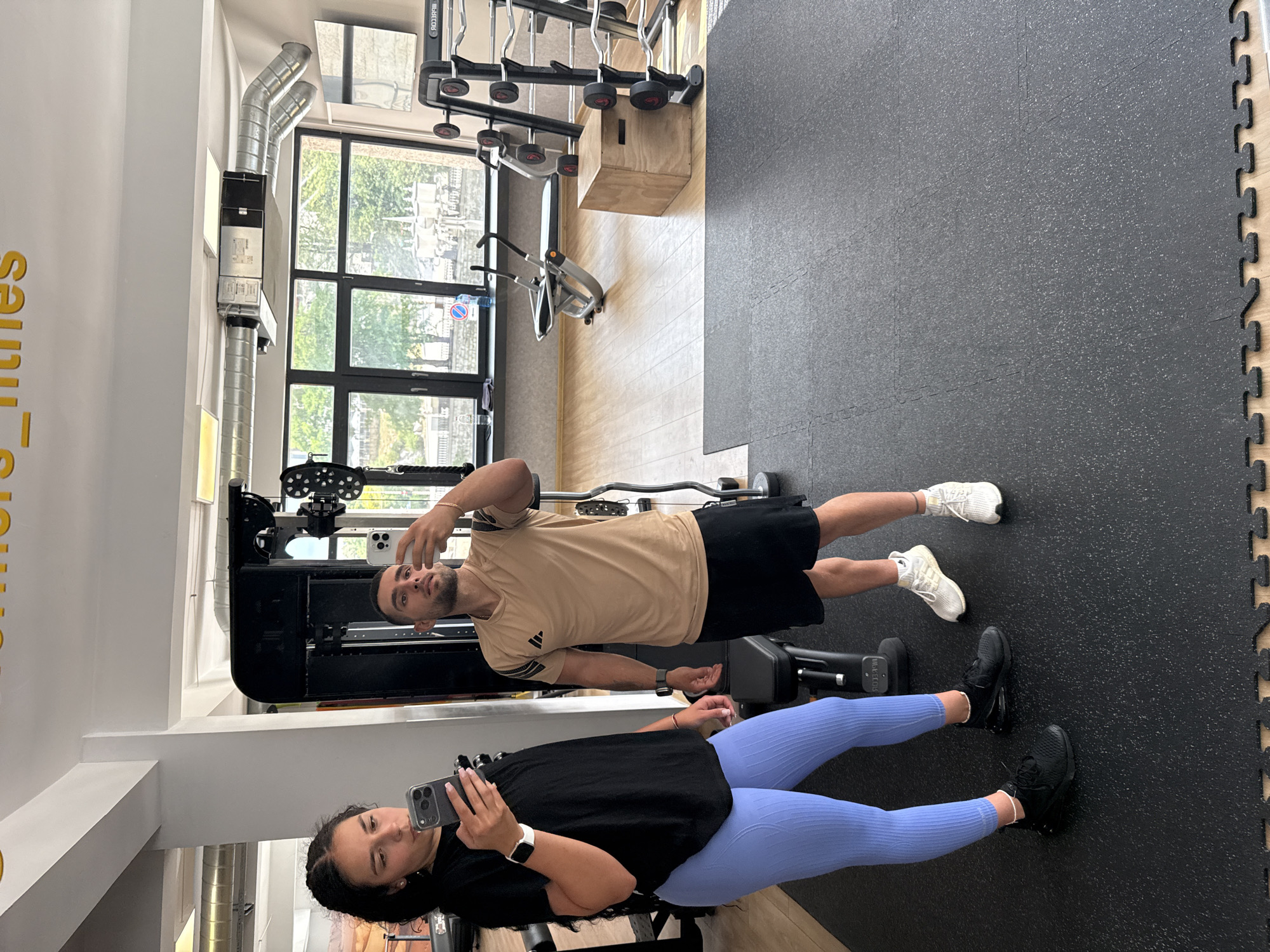 Two individuals, a woman and a man, are captured in a mirror selfie within a gym. The woman, on the left, has dark, curly hair and wears a black t-shirt and light blue leggings. She is holding a smartphone in her left hand, angled to capture the reflection. The man, to her right, has short dark hair and a beard, and is wearing a tan t-shirt with black stripes on the sleeves and black shorts. He is also holding a smartphone, taking the selfie. Both are wearing athletic shoes.
The gym environment is visible behind them. Equipment such as weight racks with barbells, a plyometric box, and other gym machines are present. Large windows in the background show trees and buildings, suggesting an urban setting. The gym floor is a combination of black rubber matting and wooden flooring. The overall scene suggests a casual workout session. The presence of the gym equipment and the windows looking out onto a street with buildings is consistent with a fitness establishment in Gabrovo, Bulgaria.