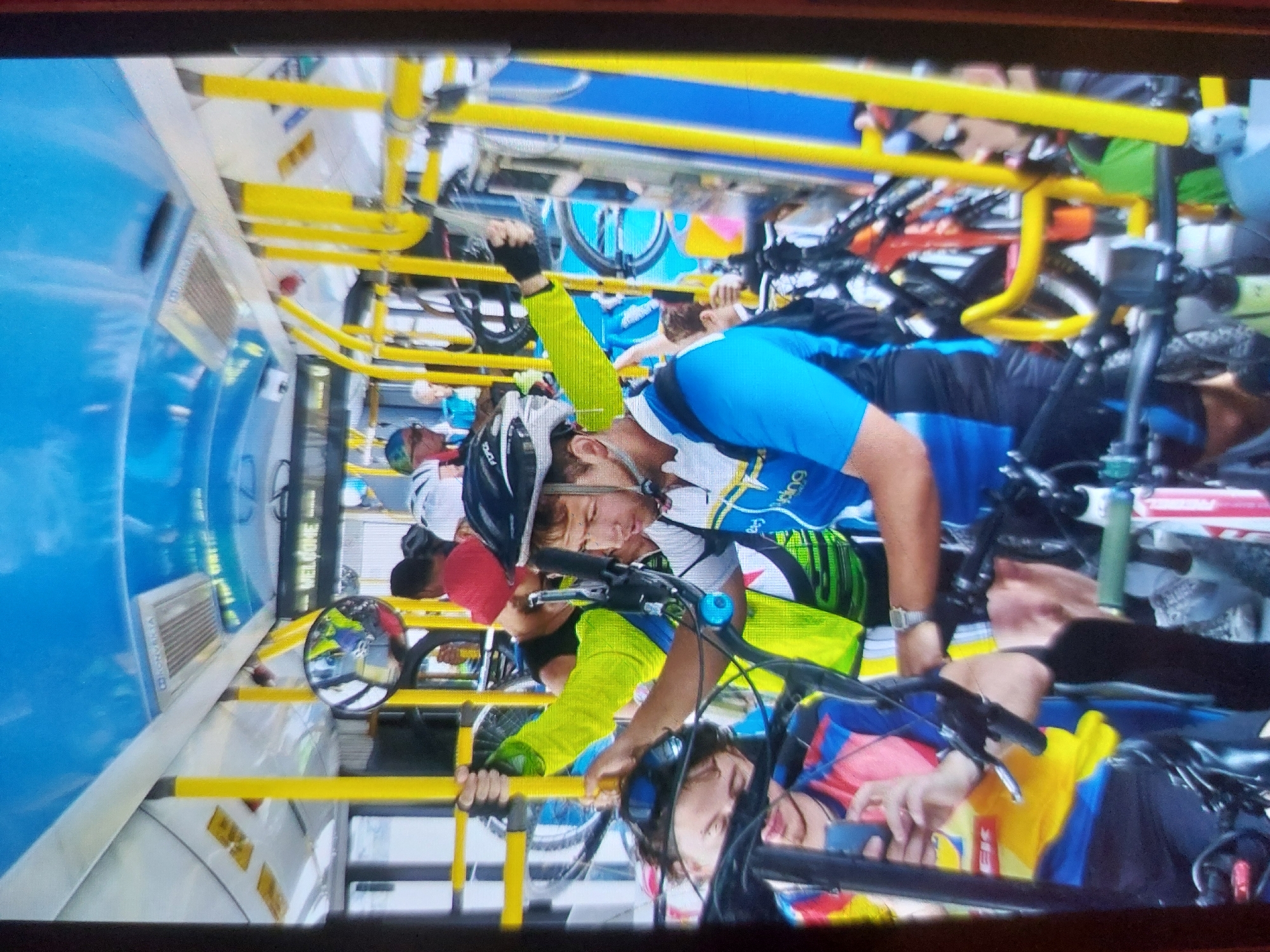 The image, rotated 90 degrees counter-clockwise, depicts the interior of a specialized bus designed for transporting cyclists and their bicycles in Tsarevo, Bulgaria. Several individuals, primarily men, are visible, clad in vibrant cycling attire including jerseys, helmets, and sunglasses. Their mountain bikes and road bikes are securely mounted vertically on bright yellow racks lining the bus interior; some bicycles appear to have their front wheels removed.
In the foreground, a man wearing a blue, white, and yellow cycling jersey, a white helmet, and sunglasses is positioned near his bike's handlebars. Below him, another cyclist's face, also helmeted, is partially visible. Further into the bus, more cyclists are seen amidst the dense arrangement of bikes. The bus interior features bright blue panels, a grey aisle, and an extensive yellow railing system for bike storage. A mirror on the left side reflects interior elements and the word "WELCOME" in reverse. A ventilation grill with the text "VOLVO" is also visible on a blue panel.
This scene captures an organized group of cyclists and their gear being transported, suggesting either the beginning or end of a cycling event or a shuttle service along a route. The overall atmosphere appears relaxed under bright daylight conditions. Additional visible text includes "600" and "GOB" on a blue jersey, and "Lidl" on a pink, red, and yellow jersey.