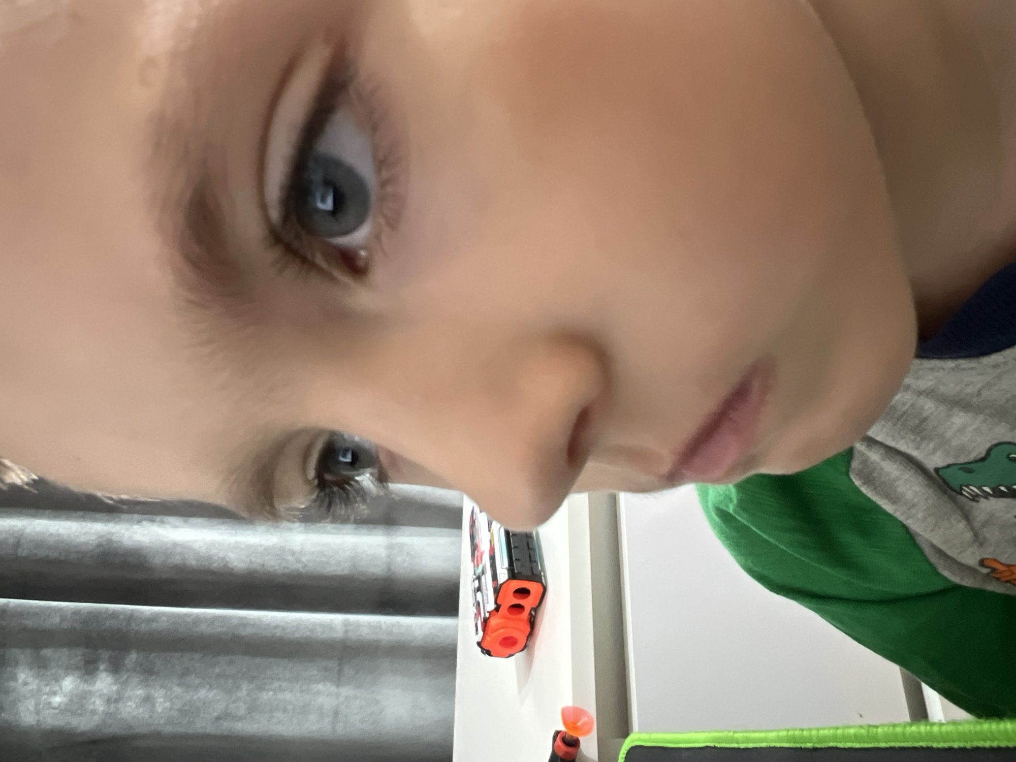A young child's face is close to the camera, angled downward. The child has light skin, blue eyes, and light hair. A Nerf gun is visible in the background, along with a gray wall and a green and gray shirt with a dinosaur design. It is not possible to determine the location of the image within the city of Марковский, Russia.