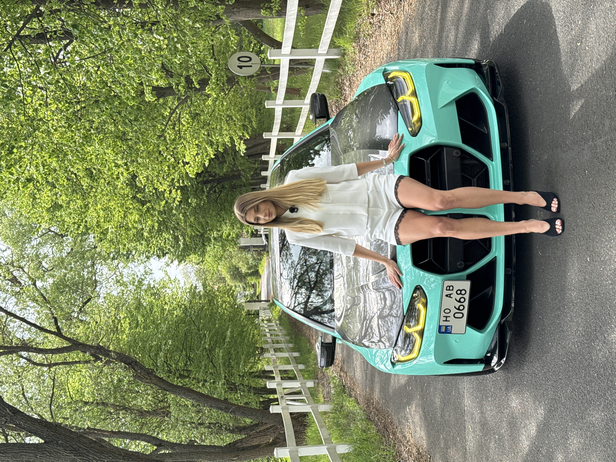 A full shot captures a blonde woman in a white ensemble reclining across the hood of a turquoise car on a road. The car has a license plate reading "HO AB 0668." A white fence runs along the left side of the road, accompanied by trees forming a leafy tunnel overhead. A sign attached to the fence displays the number "10." The location is not explicitly identified.