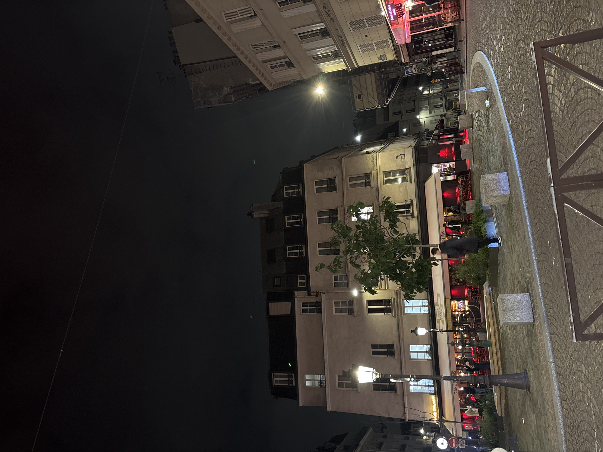 At night, the image depicts a street in Paris, France. The scene includes apartment buildings of varying heights, illuminated windows, street lamps, and an outdoor cafe setting. People are present, seated at tables enjoying the cafe, with a person walking. The street is paved with cobblestones, and there's a metal railing in the foreground.