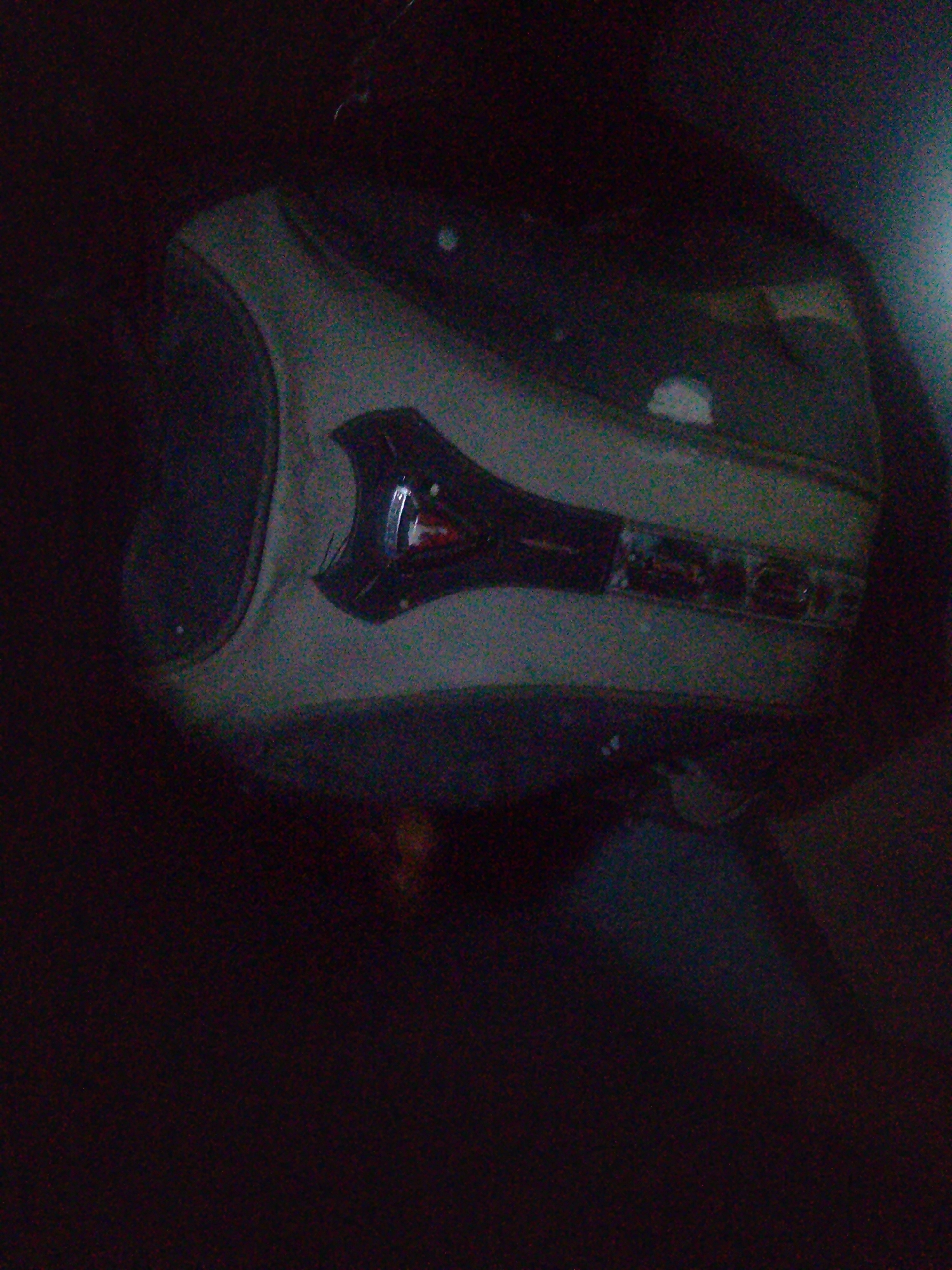 The image shows a close-up, dimly lit view of what appears to be a part of a vehicle's interior, possibly a dashboard or console. The object in focus has a curved, light-colored surface with dark accents and some reflective elements that suggest buttons or controls. A metallic piece with a red inset is visible on the left side, and a series of rectangular, dark shapes with lighter details are aligned horizontally on the right. The background is predominantly dark, with some blurred shapes and colors that do not offer clear identification of the surroundings. There is no visible indication of the city of Jalingo, Nigeria, or any context that would place the image in that location. The scene is static and appears to be an isolated detail of an object.