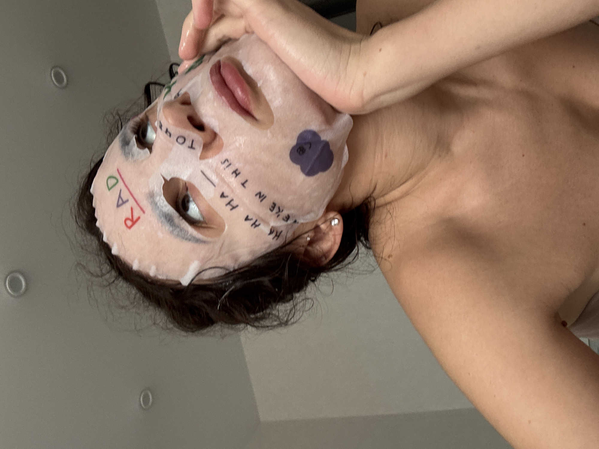 An individual, presumably in Chaikovsky, Russia, is depicted in an indoor, low-angle close-up. The person's face is covered by a translucent sheet face mask. Handwritten text in red, "RAD," is visible on the forehead, underlined. Additional text, "HA HA HA," appears on the left cheek, and "WE'RE IN THIS TOGETHER" is written across the nose and right cheek area. A purple, smiling cartoon face sticker is affixed to the lower left cheek. The individual has dark, curly hair and multiple piercings on the visible left ear, including two studs. The left hand is positioned with fingers curled under the chin, supporting the jawline. The subject is looking upwards and to the right. The upper torso is visible, suggesting bare shoulders or a strapless garment. The background consists of a plain, light-colored wall and ceiling, with two recessed circular light fixtures visible on the ceiling.