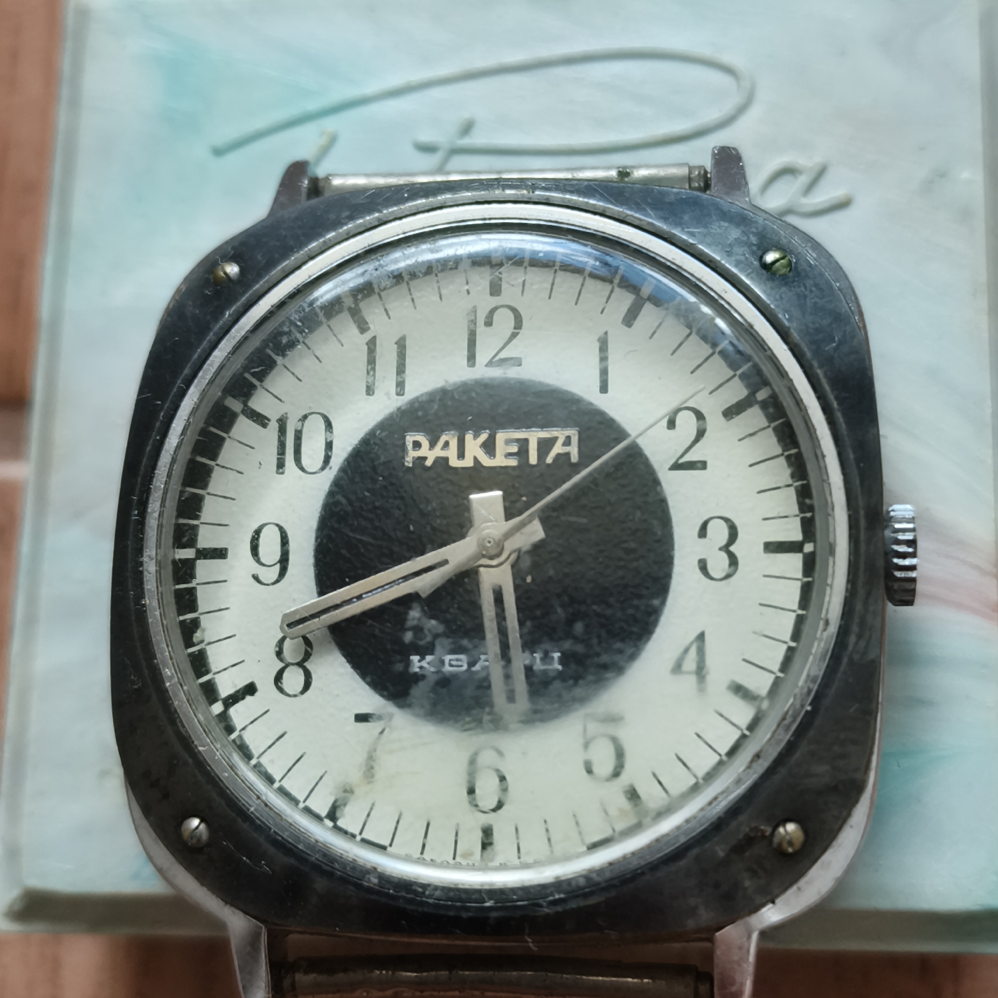 A vintage Raketa watch with a white dial and black inner circle is displayed on a light blue, textured surface. The watch features silver-toned hour markers and hands, with the brand name "RAKETA" prominently displayed on the dial. There are also Cyrillic characters below the center of the dial. The watch case appears to be made of metal and shows signs of wear. The image is taken from a close-up, slightly overhead perspective. The background is out of focus, showing a hint of a brick-like texture. There is no specific indication or context within the image that places it in the city of Chaikovsky, Russia.