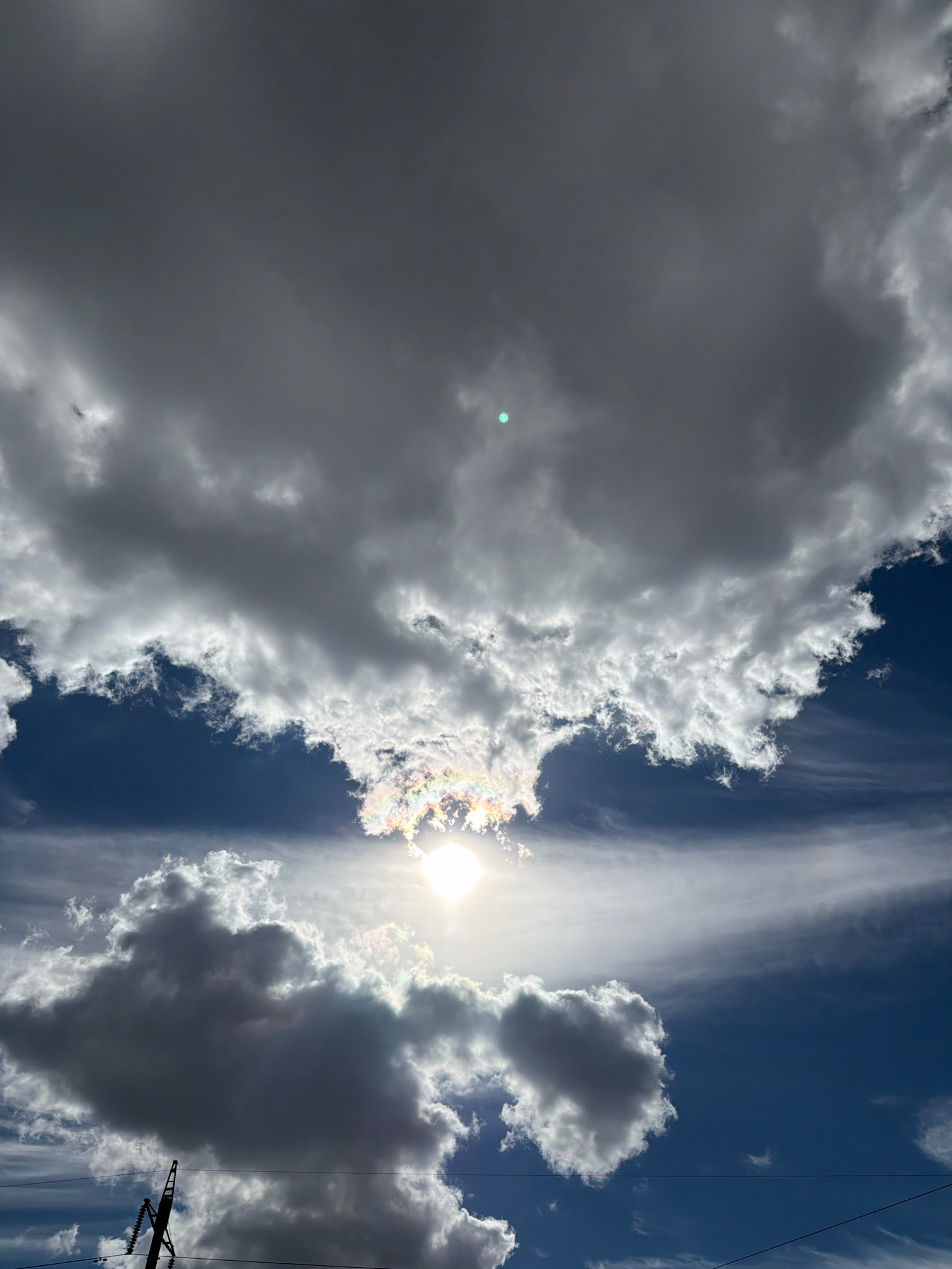 A low-angle, vertical shot captures a dynamic sky scene. The upper portion of the image is dominated by large, dark gray clouds, with lighter, wispy clouds below them. The sun is visible, partially obscured by clouds, creating a bright, white light source that also produces iridescent colors around it. The sky is a deep blue, with some streaks of white cirrus clouds present. In the lower left corner, a dark silhouette of what appears to be electrical infrastructure, including a pole and wires, is visible against the sky. There are no people or animals visible in the image. The overall impression is one of a dramatic and changing sky, possibly before or after a weather event.