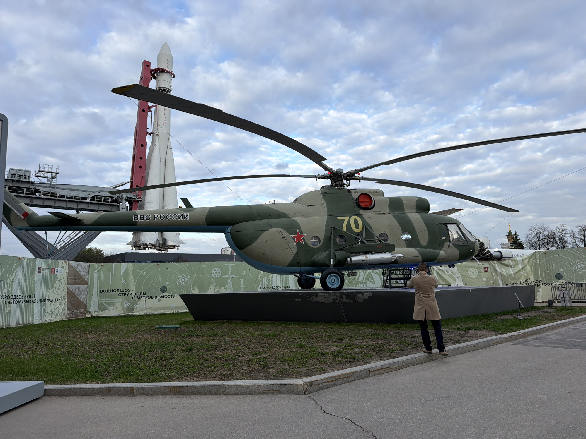 Here is a detailed description of the image:
The image shows a Mil Mi-24 attack helicopter, in a green and camouflage paint scheme, prominently displayed on a black stand at an outdoor exhibit. The helicopter bears the inscription "BBC РОССИИ" (VVS Rossii, meaning Russian Air Force) and the number "70" on its side. In the background, a tall, white rocket is visible.
A person is standing near the helicopter, facing it with their back to the camera. They are wearing a tan coat and dark pants.
The area surrounding the helicopter is well-maintained, featuring grassy patches and paved walkways. There is a fence or barrier in the background with written text on it.
The scene appears to be taking place at the Exhibition of Achievements of National Economy (VDNKh) in Moscow, Russia.