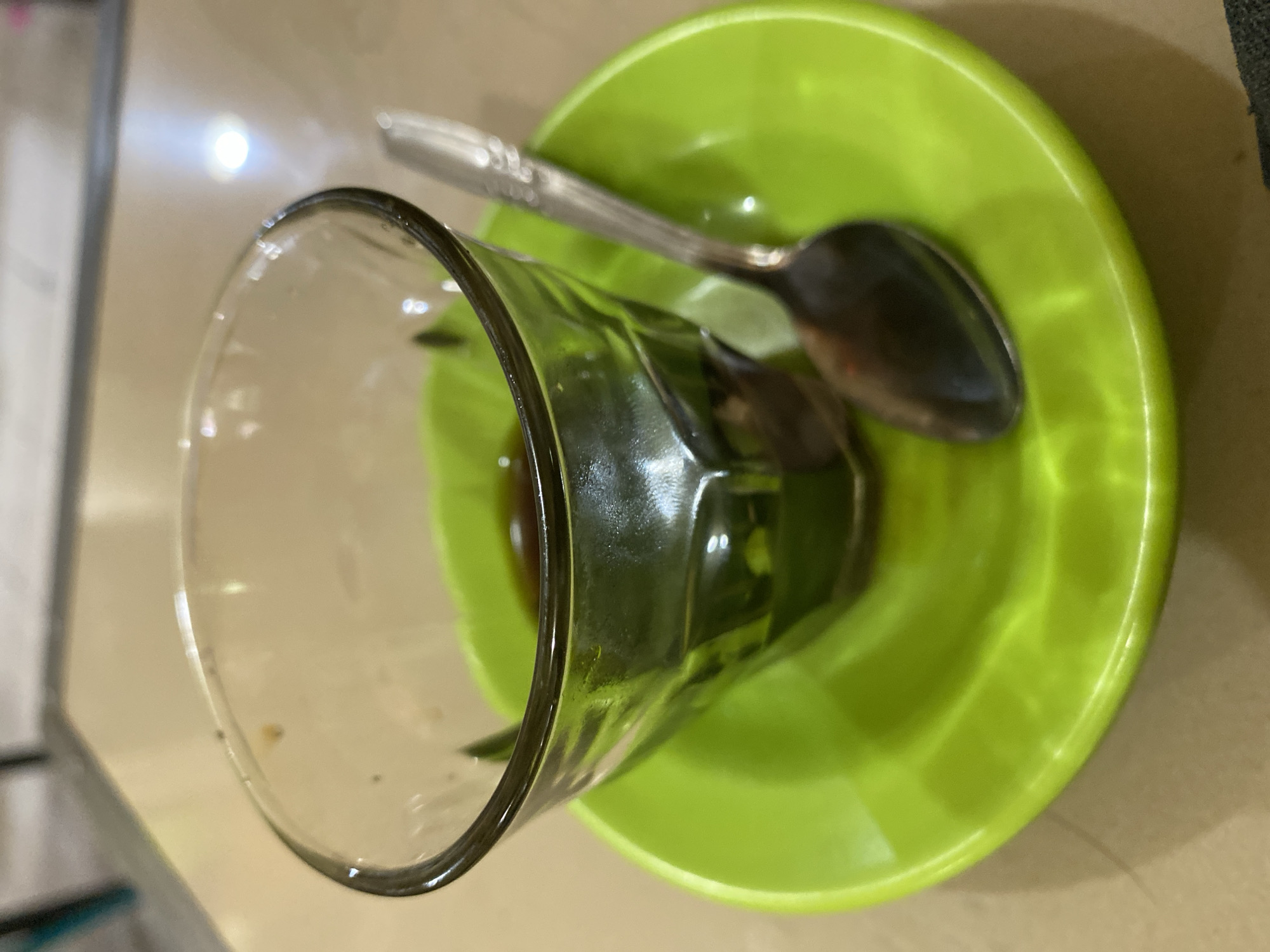 A close-up, overhead shot shows a clear glass of dark liquid, possibly coffee or tea, sitting on a bright green saucer. A silver spoon rests on the saucer to the right of the glass. The objects are placed on a beige, textured surface, likely a table. The lighting suggests an indoor setting, possibly a cafe. The image was taken in Lhokseumawe, Indonesia.