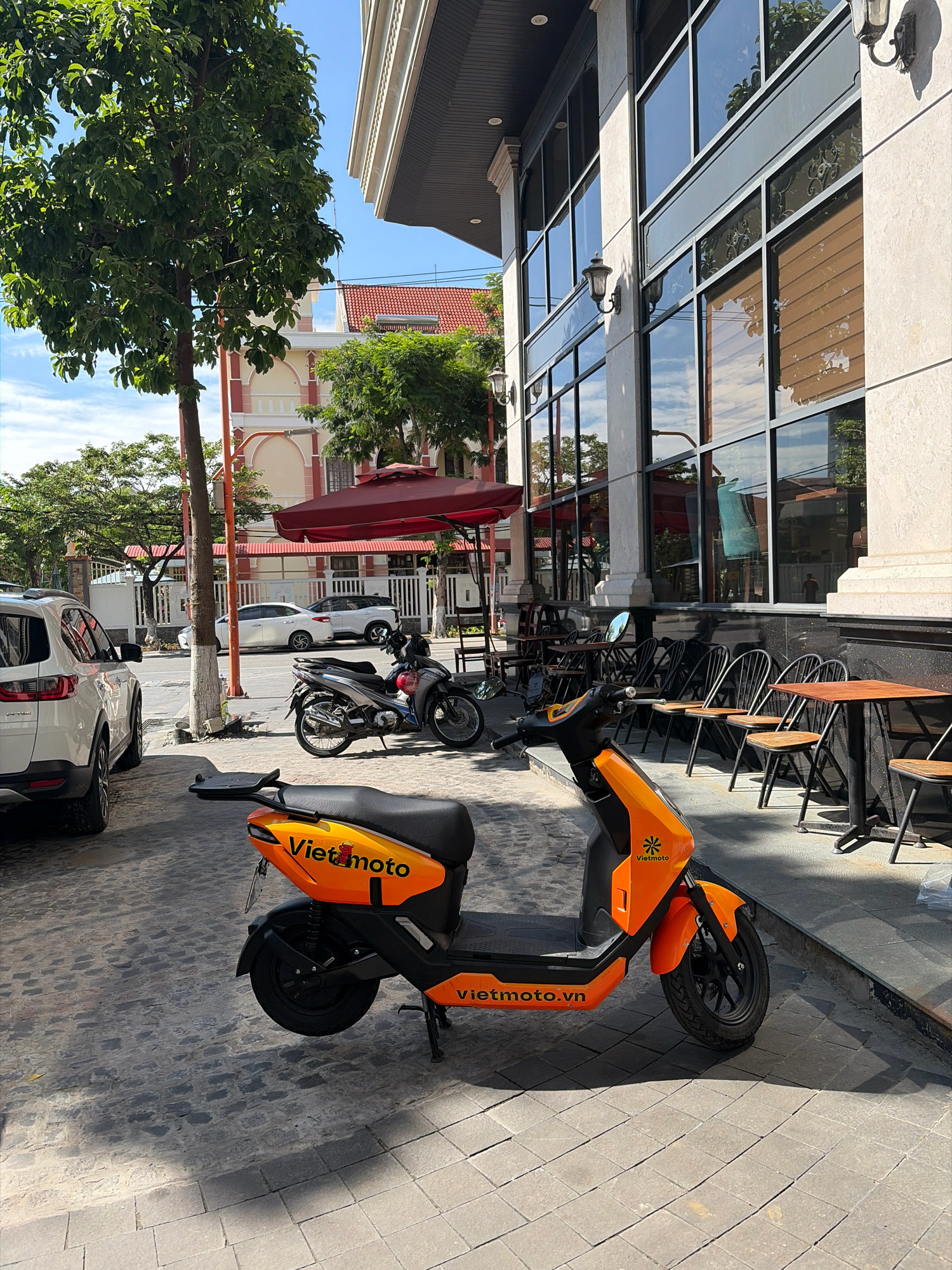 A sun-drenched street scene in Da Nang, Vietnam, showcases a vibrant orange electric scooter parked on a cobblestone sidewalk in front of a modern building with large glass windows. The scooter, emblazoned with the "Vietmoto" logo, faces the viewer at a slight angle, its black seat and orange body prominently displayed. Behind it, a silver motorcycle is parked. Further down the sidewalk, an outdoor seating area with metal chairs and tables suggests a cafe or restaurant. Across the street, cars are visible, including a white SUV and a white sedan, with a distinct pink and beige building with a red tiled roof in the background. Lush green trees and a bright blue sky with scattered clouds complete the urban landscape.