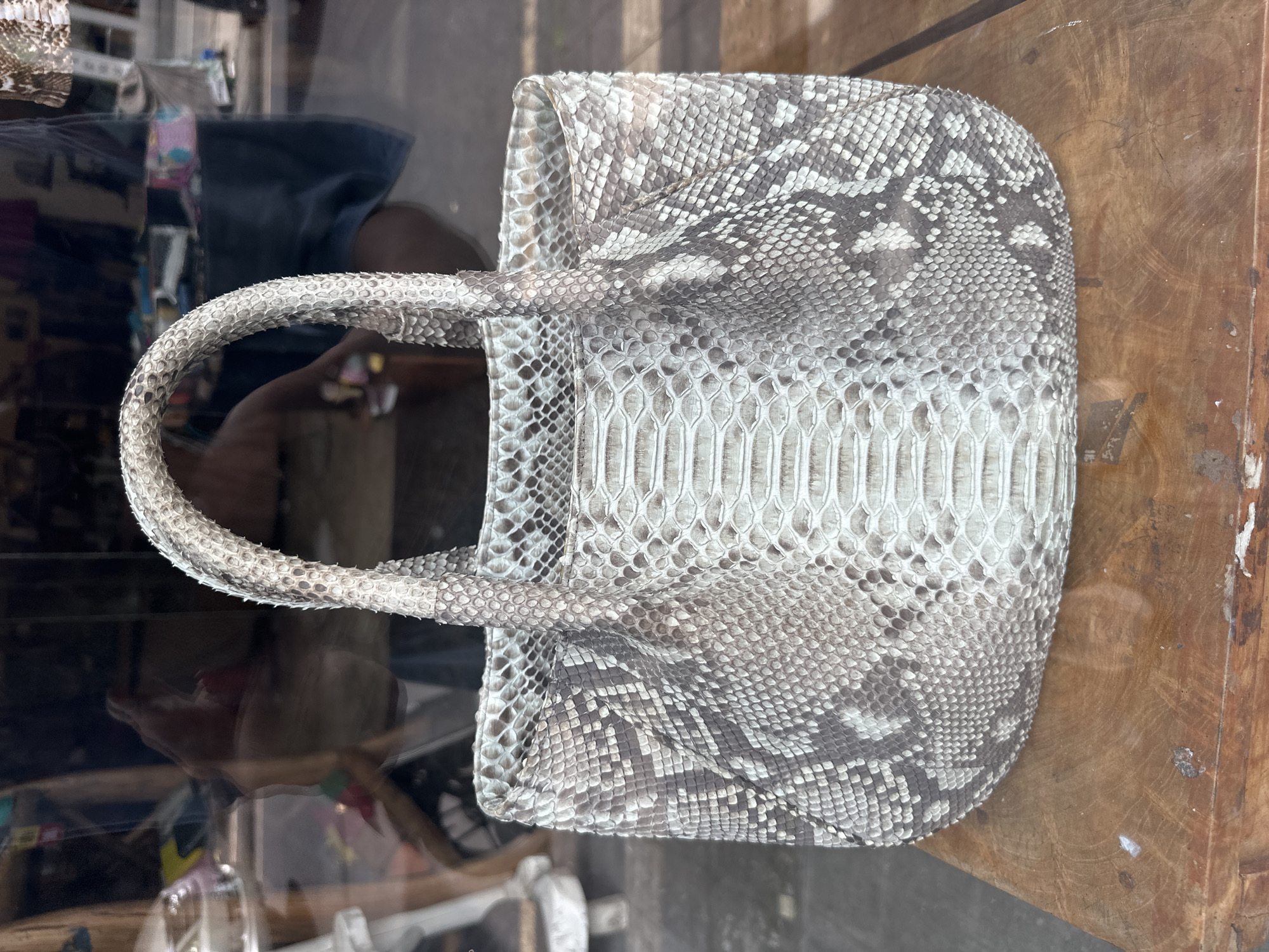 A handbag made of snakeskin rests on a wooden surface. The handbag has a textured pattern of light and dark scales and two handles. The background is blurred, showing a reflection of a person and a shop interior. The scene is outdoors, possibly in a marketplace or shop in Ubud District, Indonesia.