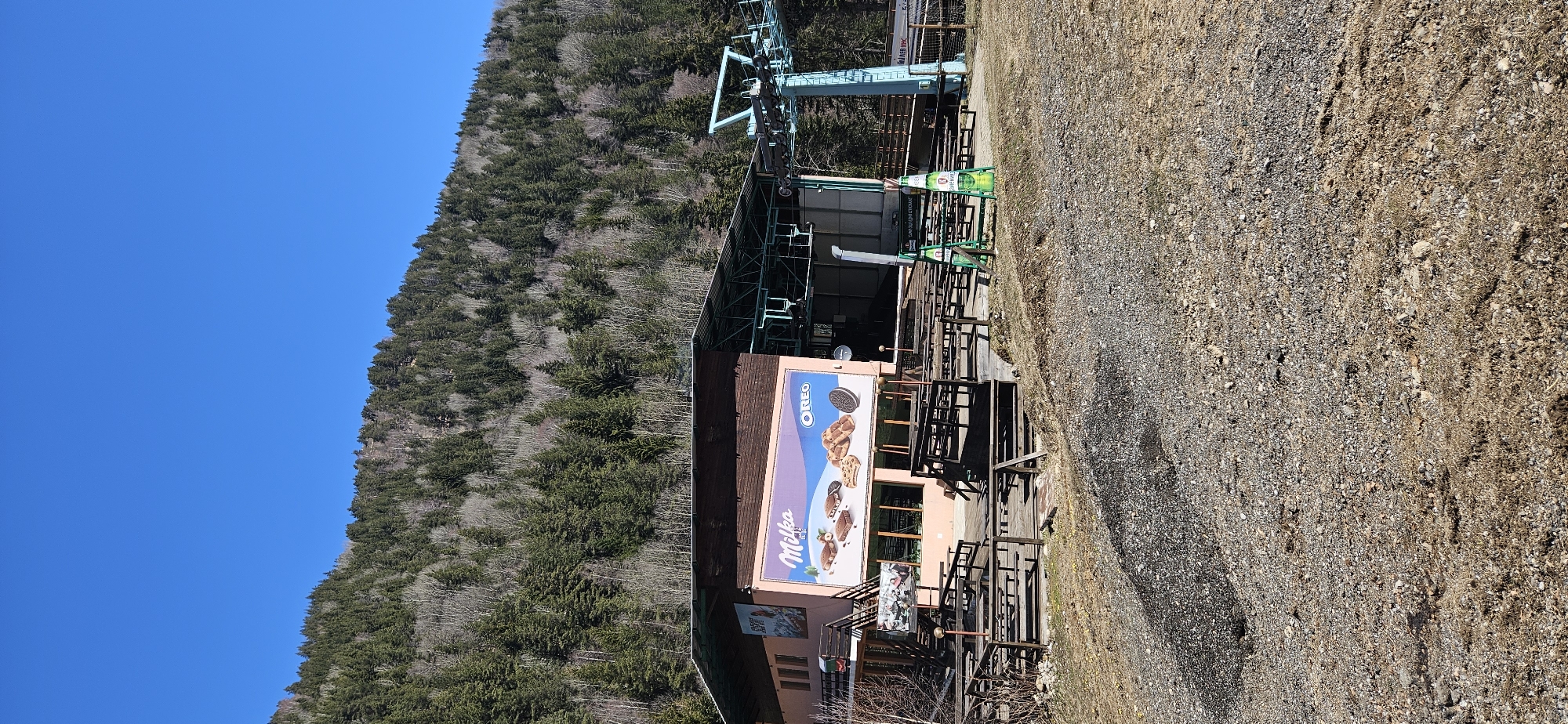 The image depicts a building with an advertisement for Milka and Oreo. The building appears to be a ski lift station or a restaurant located at a ski resort, possibly located in Blagoevgrad, Bulgaria, given the context. The station is surrounded by a forest and a rocky area. The day is clear with a blue sky. The building itself seems to have an outdoor seating area. A ski lift system is visible behind the building.