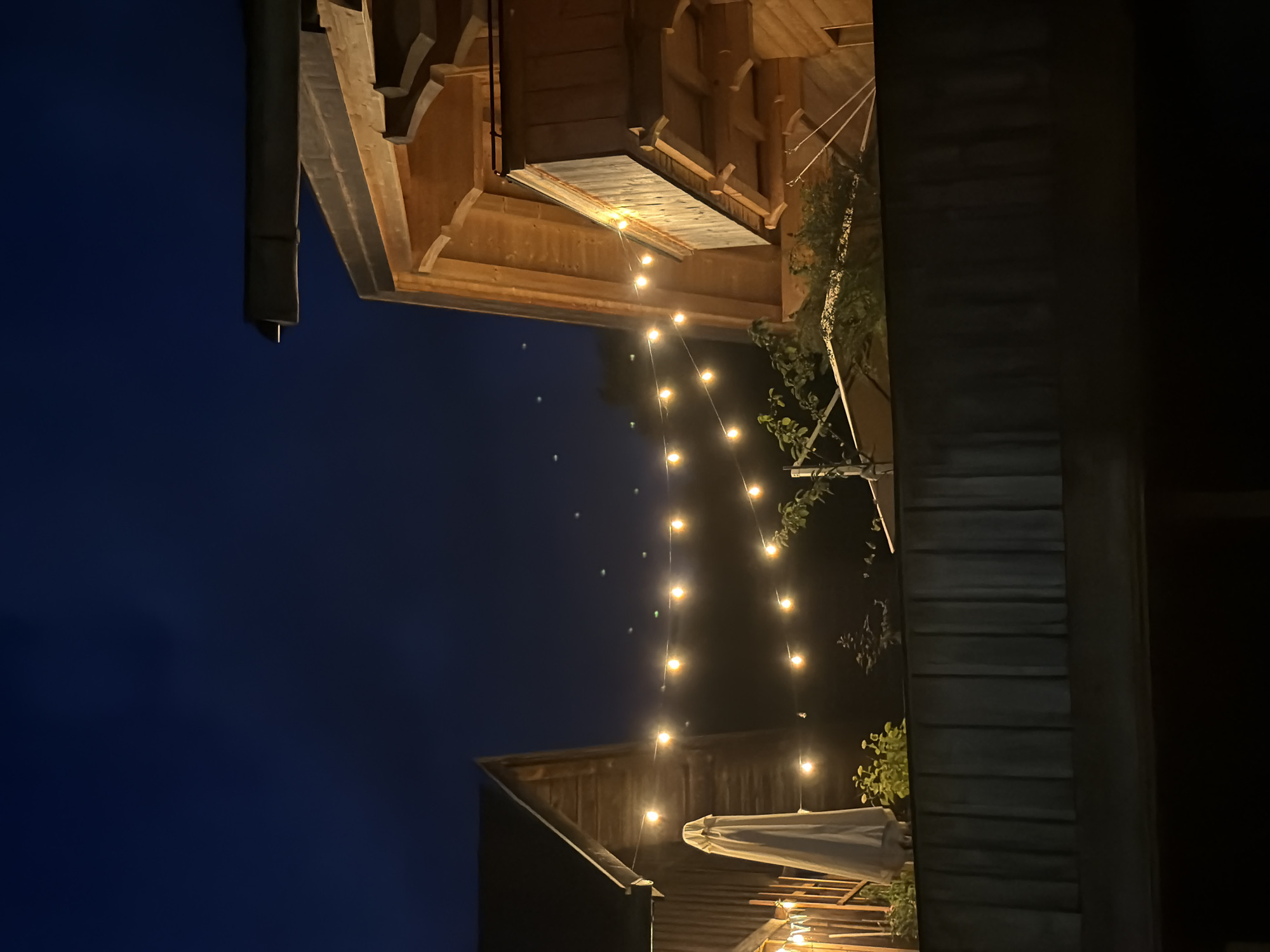 The image shows a nighttime view of two wooden buildings with string lights illuminating the outdoor area. The building on the right appears to be a multi-story structure with a balcony, while the building on the left is partially visible and seems to be a simpler wooden structure. The string lights are strung between the buildings and cast a warm glow. In the foreground, there is a dark wooden fence or railing. The sky is dark blue and clear, suggesting a pleasant evening. The scene is likely an outdoor dining or gathering area, possibly at a restaurant or a private residence. Given the architectural style and the presence of rustic wooden elements, this scene could be located in the Bavarian Alps region, and specifically Garmisch-Partenkirchen, Germany, known for its alpine charm and wooden architecture. There are no people visible in the image.