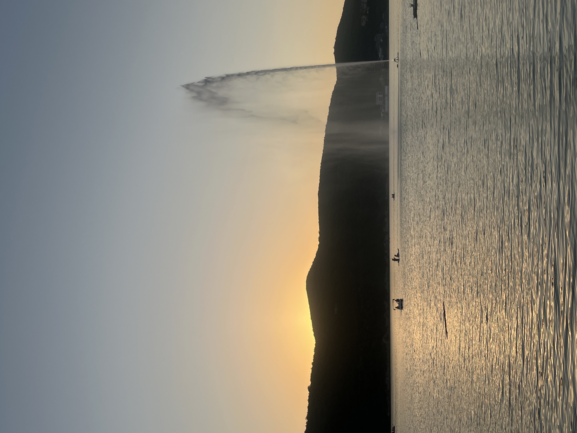 The image depicts a sunset over the Abrau Lake in Abrau-Dyurso, Russia. The sky transitions from a pale blue at the top to a warm orange and yellow near the horizon, where the sun is setting behind a silhouetted mountain range. A large fountain is prominently featured in the middle right of the lake, shooting a tall plume of water into the air. Several small boats, some with occupants, are scattered across the water's surface. The lake's surface is rippled, reflecting the warm light of the setting sun. The overall scene captures a serene and picturesque evening in Abrau-Dyurso.