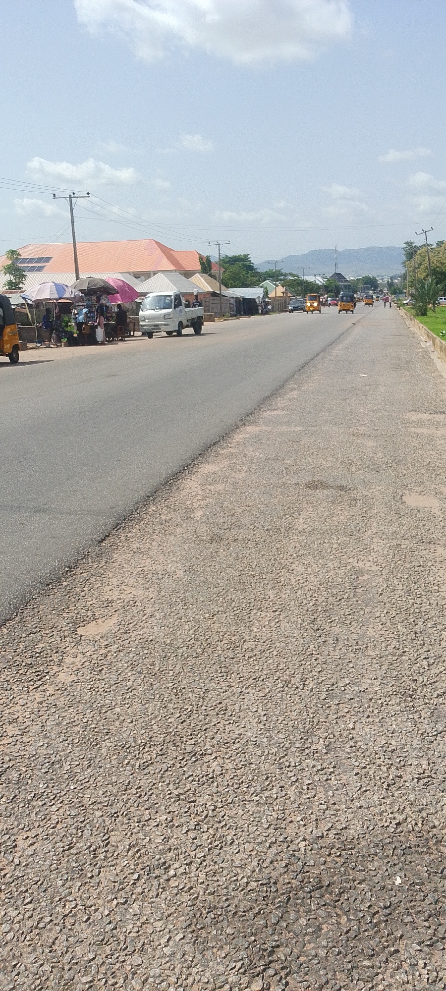 The image shows a street scene in Jalingo, Nigeria. The road is paved with asphalt in the left lane and a rougher, gravel-like material in the right lane. Along the left side of the road, there is a market area with vendors under umbrellas, selling goods. Several people are visible at the market, interacting with each other. Various vehicles are present, including a white pickup truck driving on the road, a yellow tricycle or "keke napep" behind it, and other vehicles further in the distance. Buildings with orange and gray roofs are visible behind the market stalls. Power lines and utility poles line the street. In the background, there are hills and mountains. The sky is blue with scattered white clouds, indicating it is daytime. The overall scene depicts a bustling street in a Nigerian city.
