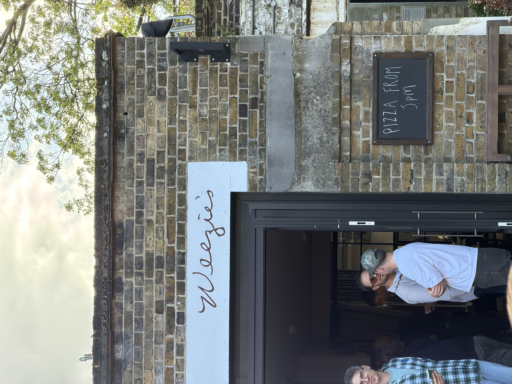 The image depicts the exterior of "Weezie's," a business in London, United Kingdom. The building is constructed of light brown bricks. A sign next to the entrance reads "Weezie's" in brown cursive font on a white background. A chalkboard sign on the brick wall says "PIZZA FROM 5pm." Three people are partially visible in the doorway. One is wearing a white shirt and a camouflage baseball cap and appears to be talking with another individual who is wearing sunglasses. A third person wearing a blue and green plaid shirt and glasses is partially visible at the bottom of the image. The weather appears to be sunny. A tree is visible in the upper left corner.