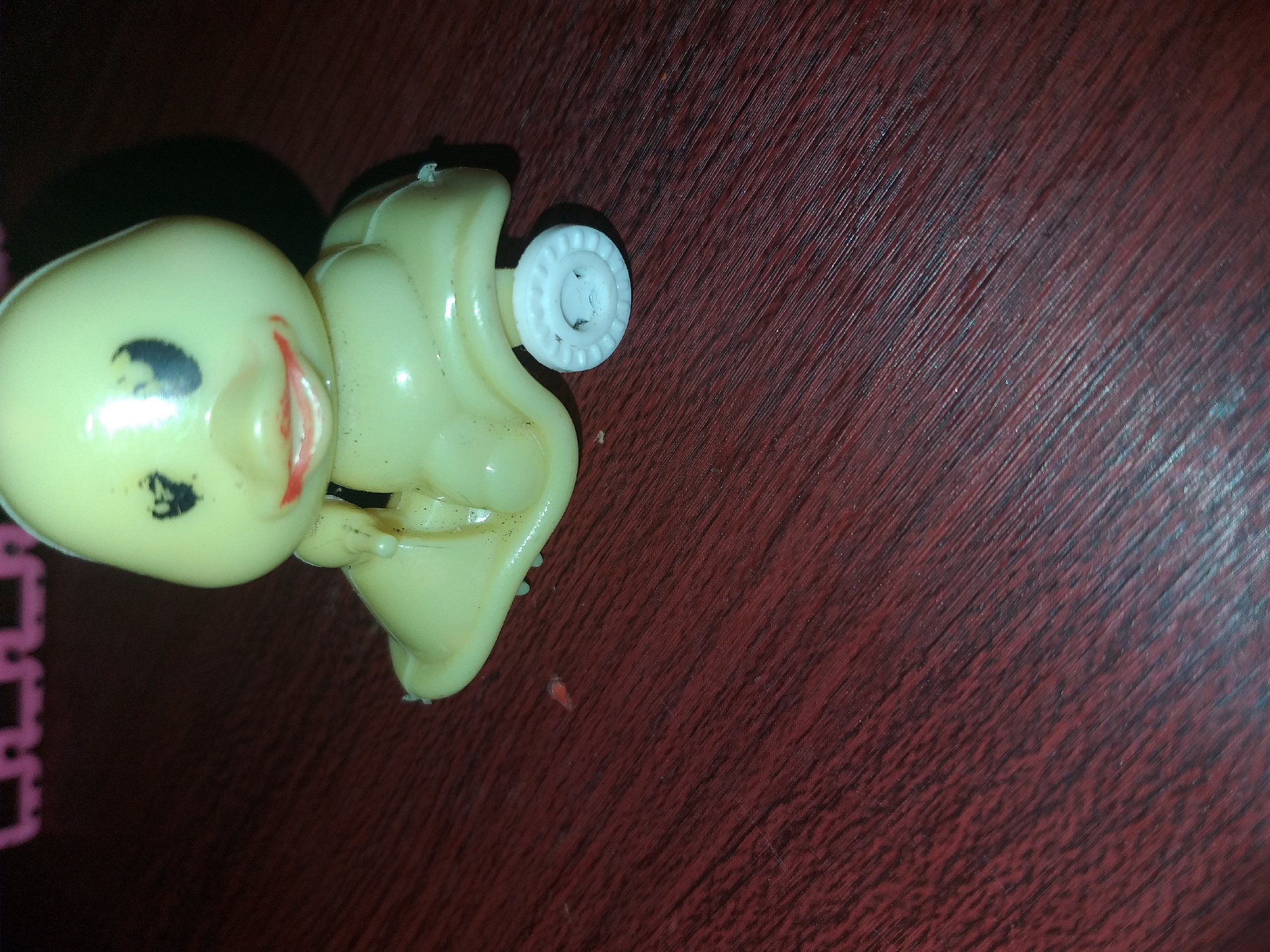 A close-up shot, likely taken indoors, shows a light yellow plastic duck toy with black eyes and red lips, mounted on a white wheel. The toy appears to be a pull-along toy, designed to move on wheels. It is resting on a dark red, wood-grain surface, possibly a table or floor. In the background, out of focus, a row of pink plastic objects is visible. The lighting is dim, casting a shadow to the right of the duck. There are no people or animals present in the image. The location in Air Jamban, Indonesia, is not discernible from the visual information provided in the image.