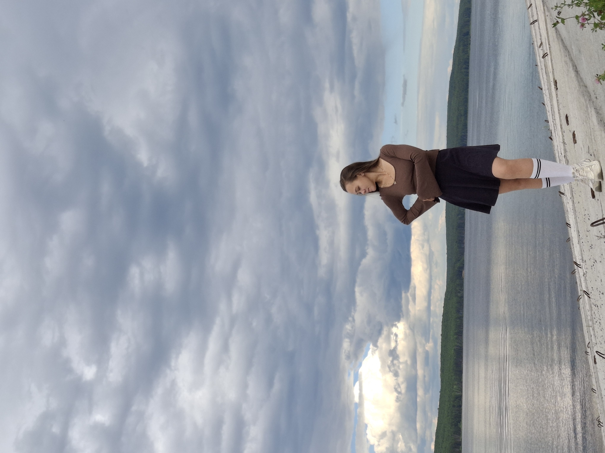 The media file is a full-body, low-angle, sideways portrait of a young woman standing on a concrete structure overlooking a large body of water. She is wearing a long-sleeved brown top, a dark skirt, white socks with black stripes, and white sneakers. Her hair is dark and worn down. She is looking downwards, with a thoughtful or contemplative expression.
The scene is set outdoors with a dramatic sky filled with clouds. The sky transitions from a lighter, brighter area on the right to darker, more overcast clouds on the left. A vast body of water stretches across the background, with a distant, tree-lined shore visible on the left side. The concrete structure the woman is standing on appears to be a walkway or pier, with visible metal elements and a textured surface. Small pink flowers are growing on the edge of the structure on the far right.
The overall mood is calm and serene, possibly evoking a sense of introspection. The lighting suggests it is either early morning or late afternoon, with the sun breaking through the clouds in some areas. The location cues, like the specific type of vegetation and the vastness of the water, combined with the provided context of Chaikovsky, Russia, suggest a natural landscape setting. There is no visible text in the image.