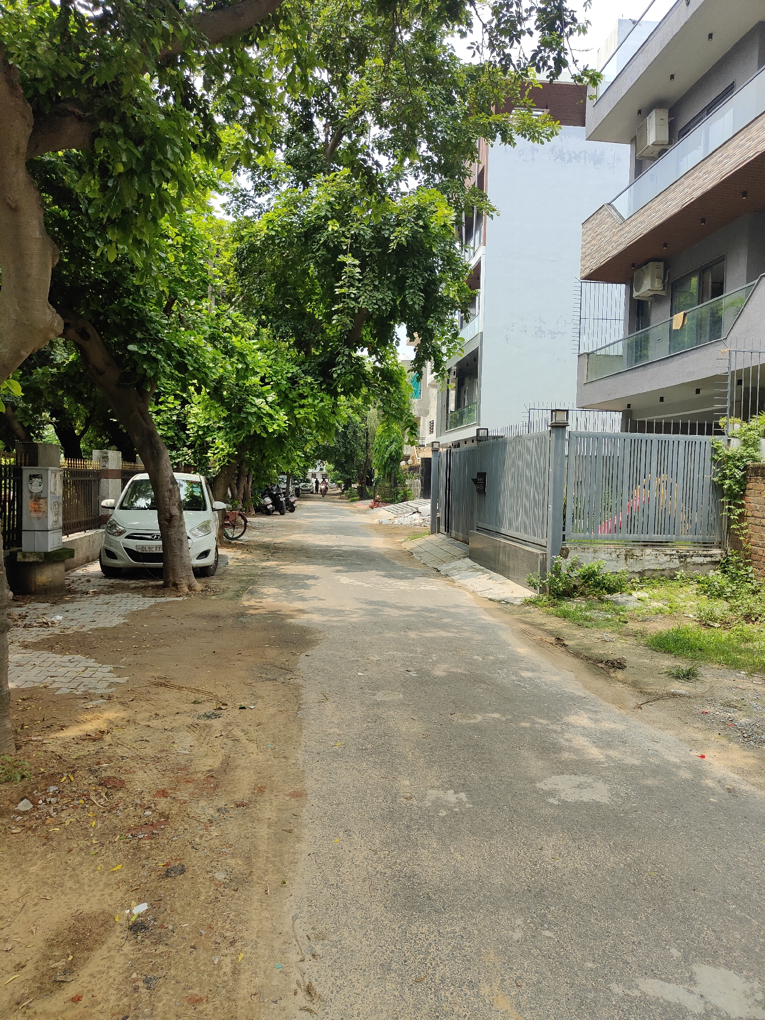 The image shows a street in Gurgaon, India, bordered by trees and modern buildings. On the left side of the street, a white car is parked next to a sidewalk made of patterned bricks. A tree trunk is visible in the foreground, partially obscuring the car. A metal fence runs along the sidewalk, with an electrical meter box mounted on a concrete pillar next to it. Further down the street, there are parked motorcycles and a bicycle. On the right side of the street, a contemporary building with multiple balconies and glass railings is visible, with a grey metal gate in front of it. The street itself is paved with asphalt and appears to be well-maintained, though there are patches of dirt and debris on the sides. The environment is urban, with evidence of both residential and commercial activity. The overall impression is one of a quiet, leafy residential street in a modern Indian city.