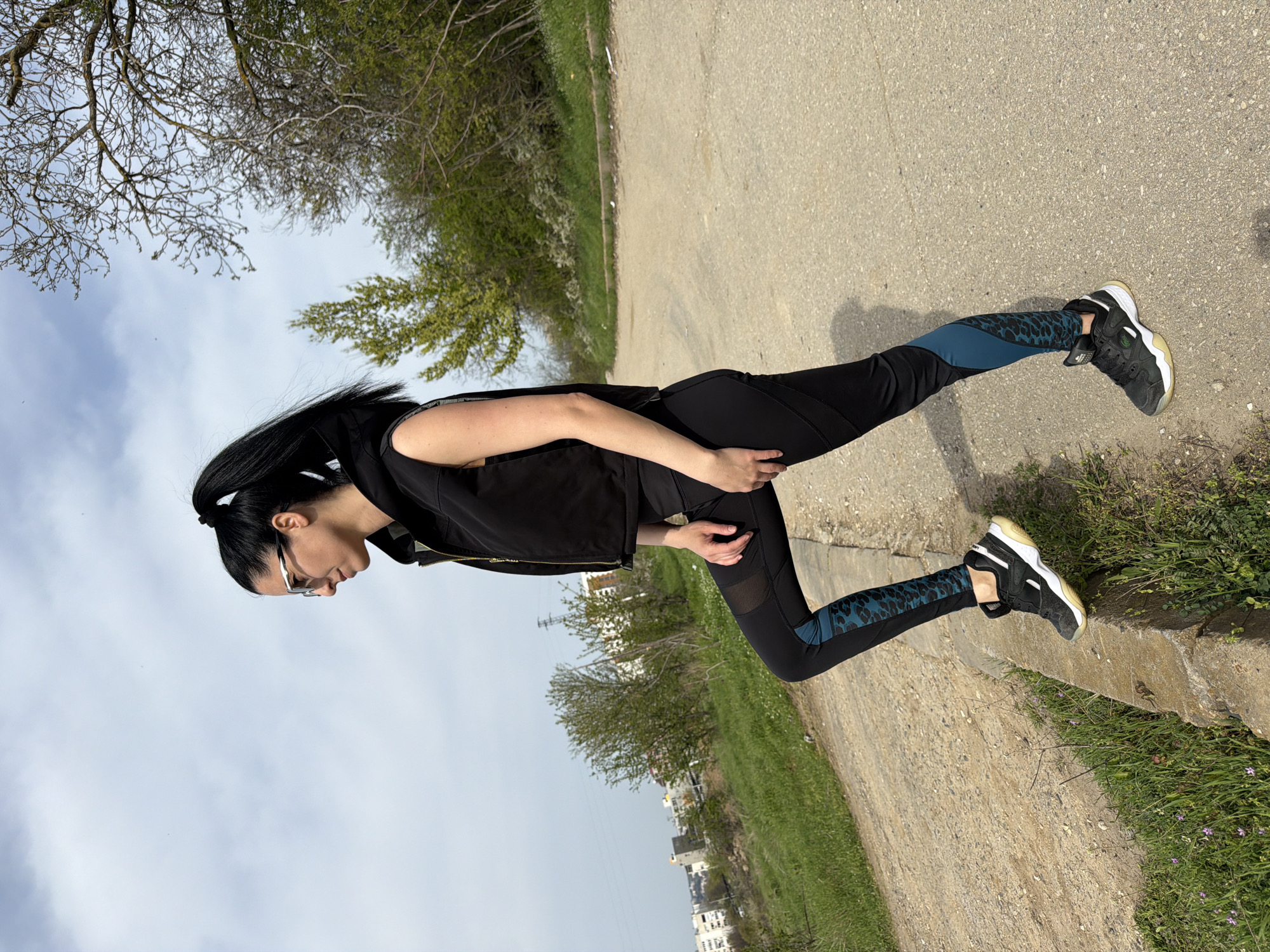 A woman stands on a concrete path next to a stone wall in Pleven, Bulgaria. She is wearing black leggings with teal and animal print panels, a black vest, and black and white sneakers. Her hair is pulled back in a ponytail, and she is wearing glasses. In the background, there is vegetation and a cloudy sky. Across the path are trees and buildings.