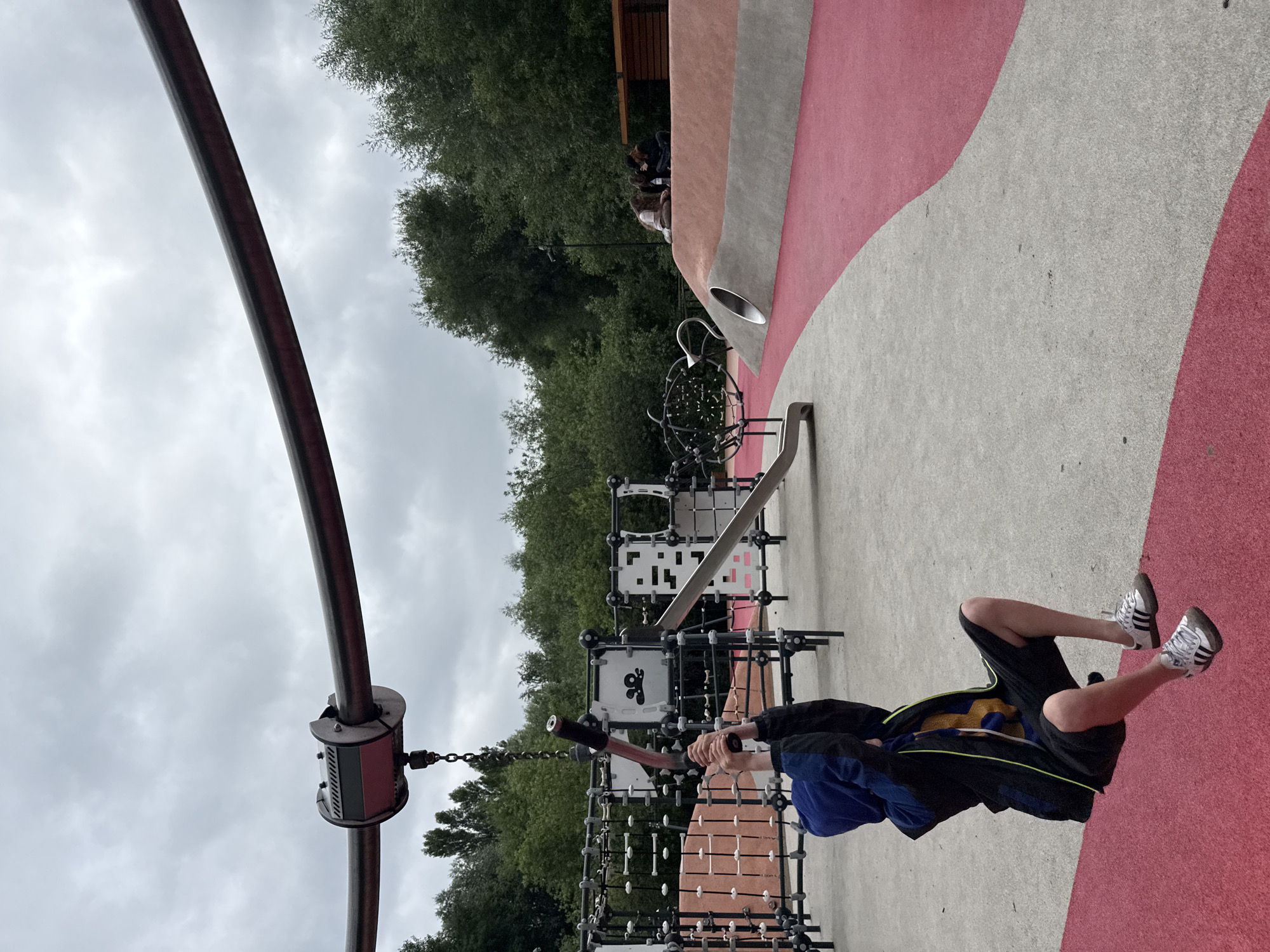 A boy is riding a zip line in a playground in Moscow, Russia. The playground features a grey and red rubberized surface with various climbing structures and slides. The boy, wearing a blue hooded jacket and black shorts, is holding onto the zip line with his hands. In the background, several people are sitting on a raised embankment, and lush green trees form the backdrop under a cloudy sky.