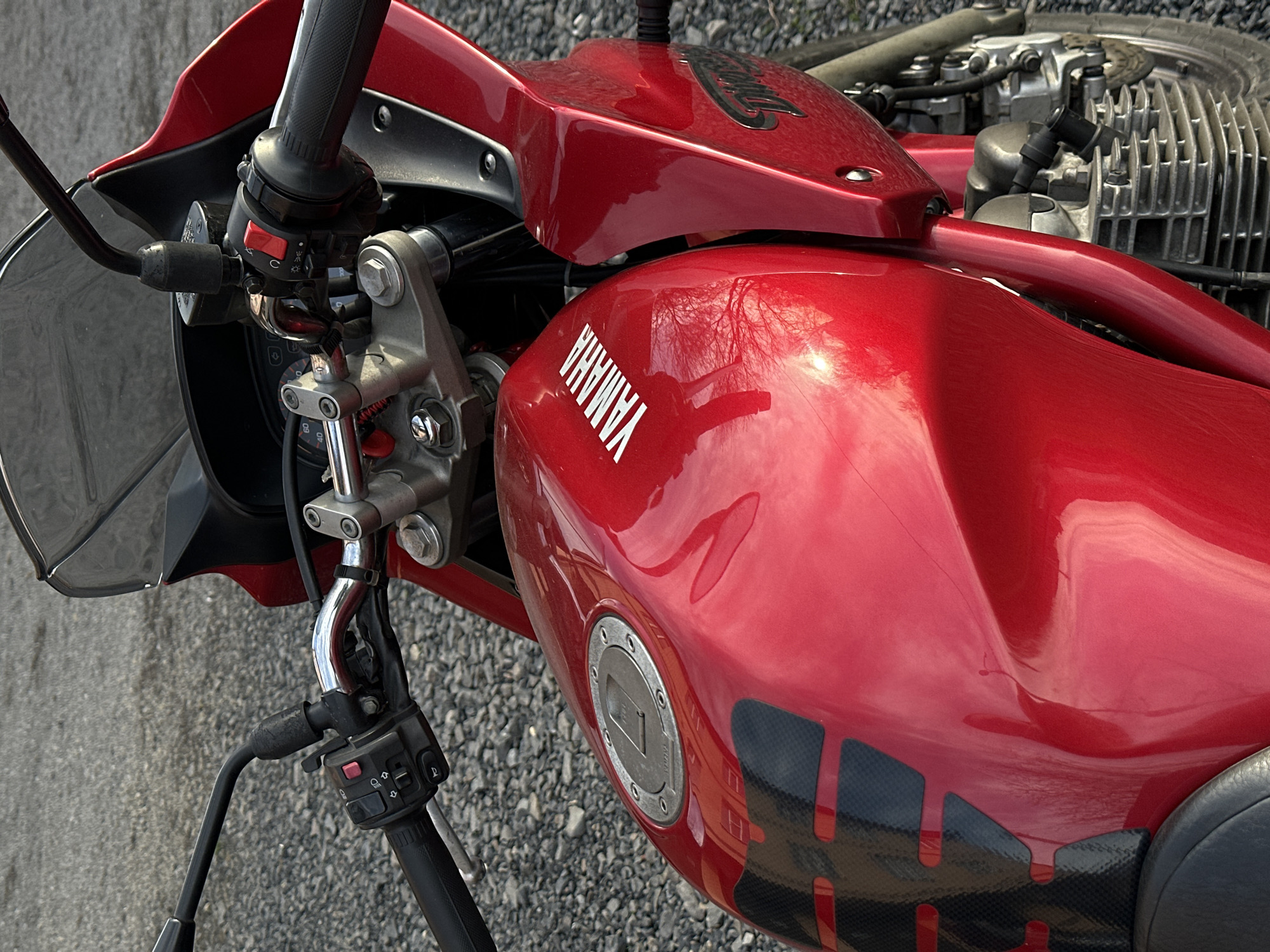 A close-up shot captures the front of a red Yamaha motorcycle. The motorcycle is parked on a gray gravel surface. The handlebar and its controls are visible on the left, along with a small windshield. The fuel tank is prominent, featuring the Yamaha logo. The gas cap is silver. There are black decals present on the fuel tank. The motorcycle's engine is partially visible on the right side of the frame.
There is no information available in the image to suggest the location. Therefore, the city of Rostov-on-Don, Russia cannot be confirmed.