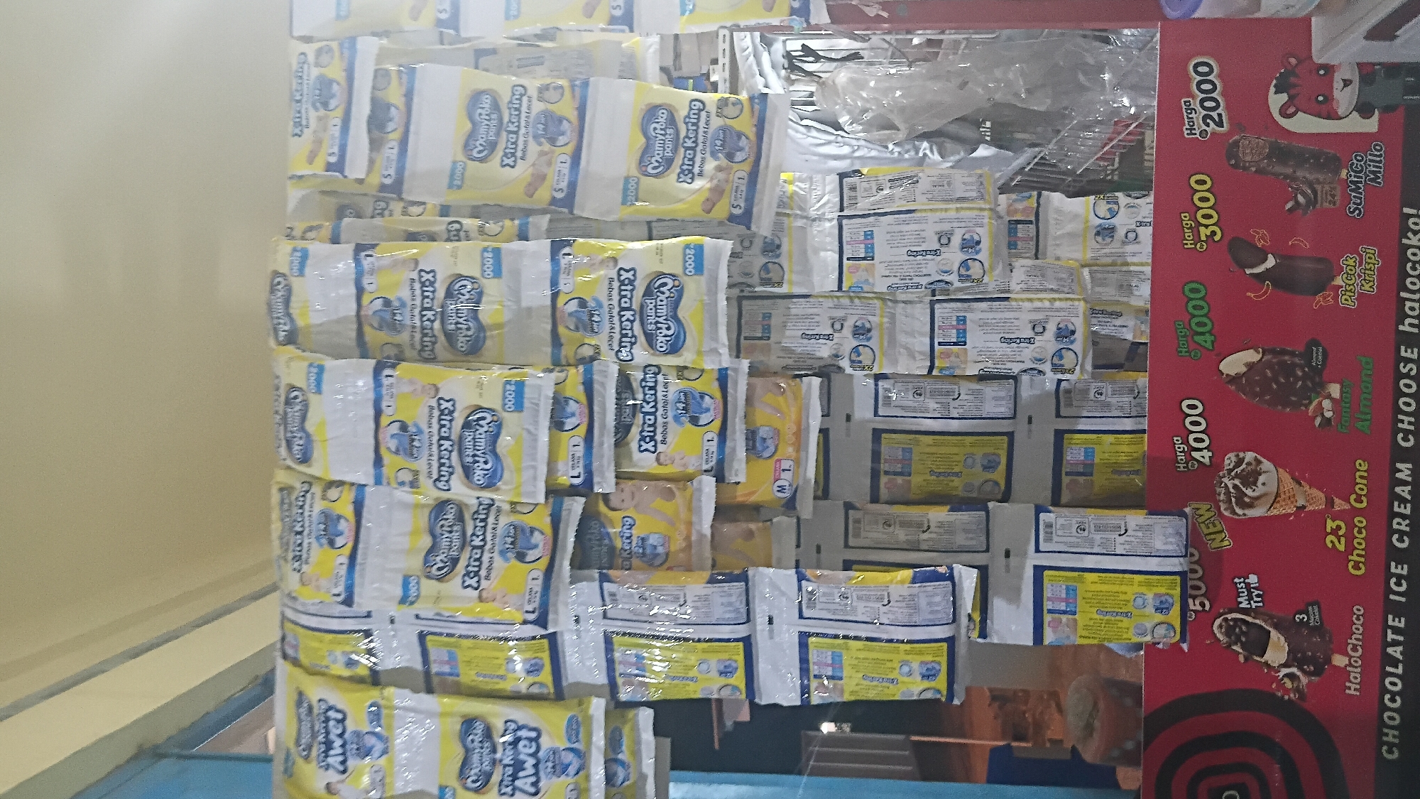This image captures a retail display in Prabumulih, Indonesia, likely at a small convenience store or warung. The primary focus is a wall covered with numerous packs of baby diapers, predominantly branded "MamyPoko Pants Xtra Kering". These packs are hung in rows, creating a dense visual texture.
Below the diaper display, a vibrant red sign advertises various ice cream products. The sign displays prices in Indonesian Rupiah (Rp) and features illustrations of different ice cream cones and bars. Specific ice cream names visible include "Halo Choco", "Choco Cone", "Fantasy Almond", "Piscok Krispi", and "SuMico Millo". A cartoonish character with a tiger-like face, wearing overalls, is also depicted on the right side of the ice cream advertisement.
The background shows a cream-colored wall and a glimpse of a blue painted structure to the left. The lighting suggests an indoor setting, possibly with artificial lights, as there are no strong shadows indicative of direct sunlight. The overall impression is one of a well-stocked, local retail environment catering to everyday household needs and small treats.
