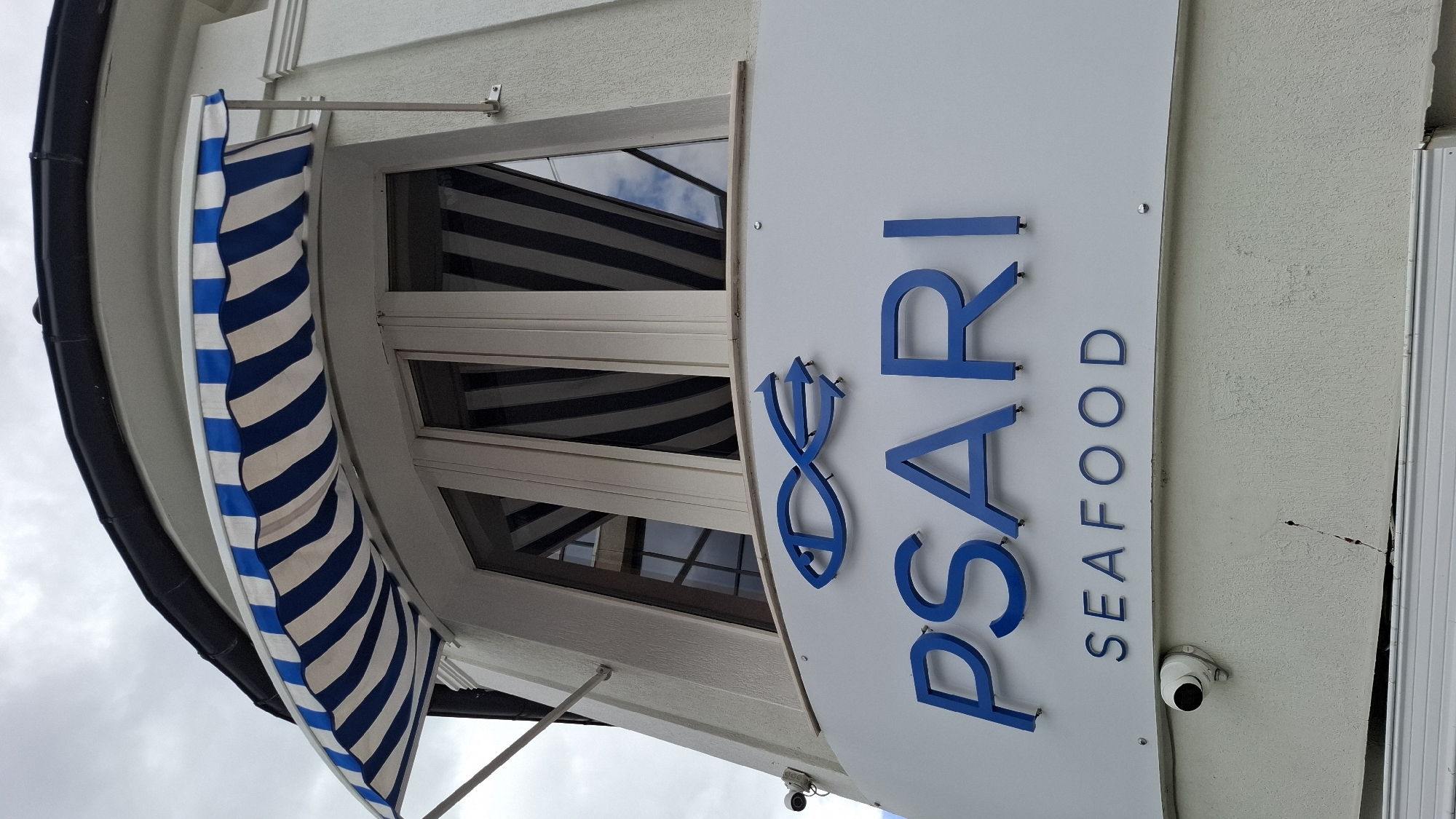 The image shows the exterior of a seafood restaurant named "PSARI SEAFOOD" located in Sofia, Bulgaria. The building features a white facade with a blue and white striped awning above a set of windows. The restaurant's name is displayed in large blue letters, with a stylized blue fish symbol above it. The word "SEAFOOD" is written in smaller blue letters below the restaurant's name. There are two security cameras visible on the building's exterior, one on the upper level and one on the lower level. The sky in the background is overcast.