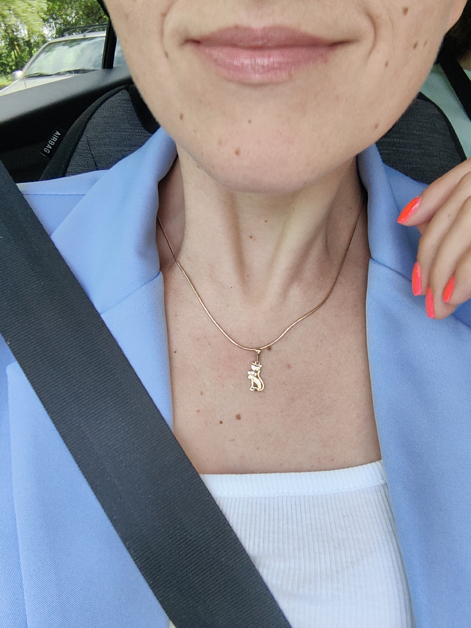 The image depicts a close-up, chest-up view of an individual seated in the passenger compartment of a vehicle, in Дабрынёўскі сельскі Савет, Belarus. The subject's light-toned skin displays scattered moles and freckles across the chin, jawline, and upper chest. A faint smile is indicated by a crease on the viewer's right cheek and slightly parted, pink lips. The individual wears a light blue blazer over a white ribbed tank top. A dark grey seatbelt diagonally crosses the chest from the upper left to the lower right. A thin, gold-colored snake chain necklace with a small, stylized gold cat pendant is worn around the neck. The left hand (viewer's right) is visible near the shoulder, with all fingernails painted a vibrant neon coral. The vehicle's interior features a dark grey fabric seat and a visible "AIRBAG" label on the side panel. Through the car window in the upper left background, green foliage and part of a light-colored SUV with roof racks are discernable, indicating an outdoor, daytime setting.