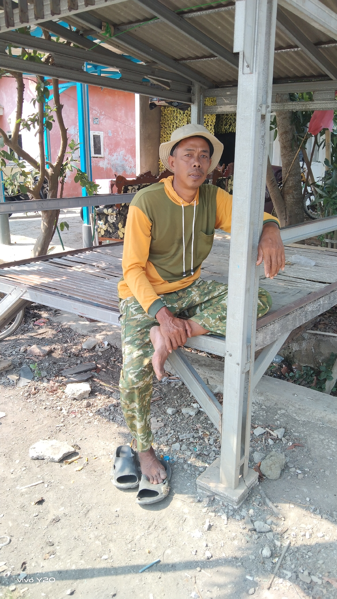 A man in a light-colored bucket hat and a long-sleeved shirt, split between olive green and yellow, is seated on a weathered, slatted metal platform. He wears camouflage pants and black slide sandals. His left foot rests on his right knee, and his hands are clasped.
The setting appears to be an outdoor, partially covered area. A metal structure with a corrugated roof arches overhead, supported by gray metal poles. Behind the man, a pink building with blue trim is visible, along with trees. The ground is a mix of dirt, gravel, and scattered debris. Sunlight indicates it is daytime.
The man's expression is neutral as he looks towards the camera. The scene suggests a casual, perhaps resting moment, in an outdoor environment that could be a rural or semi-urban setting in Karawang, Indonesia. The "VIVO Y20" watermark is visible in the bottom left corner.