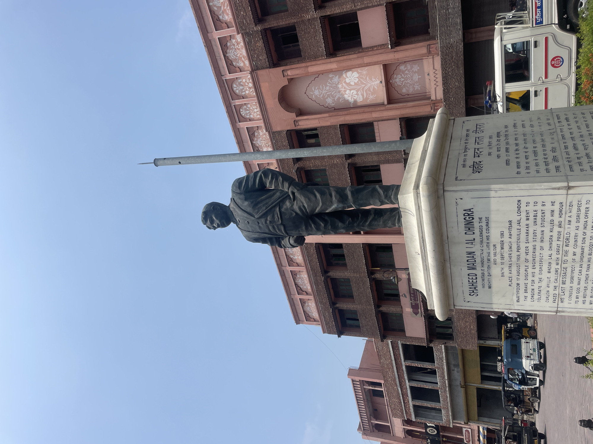 The photograph captures an outdoor scene in Amritsar, India, centered on a statue of Shaheed Madan Lal Dhingra. The statue is positioned on a large white pedestal with inscriptions in English and what appears to be Punjabi. Dhingra is depicted as a man in a suit, standing with his hands in his pockets, facing forward.
Behind the statue, a large, ornate building with a pink facade is visible. This building features architectural details such as arched windows and decorative floral motifs. To the left of the statue, another building with a reddish-brown brick exterior and arched openings can be seen. The sky is clear and blue, indicating a sunny day.
In the foreground and to the left of the statue, several auto-rickshaws are parked, and a white police vehicle with blue flashing lights is present to the right of the pedestal. The ground appears to be paved. The overall atmosphere is that of a public square or monument area.