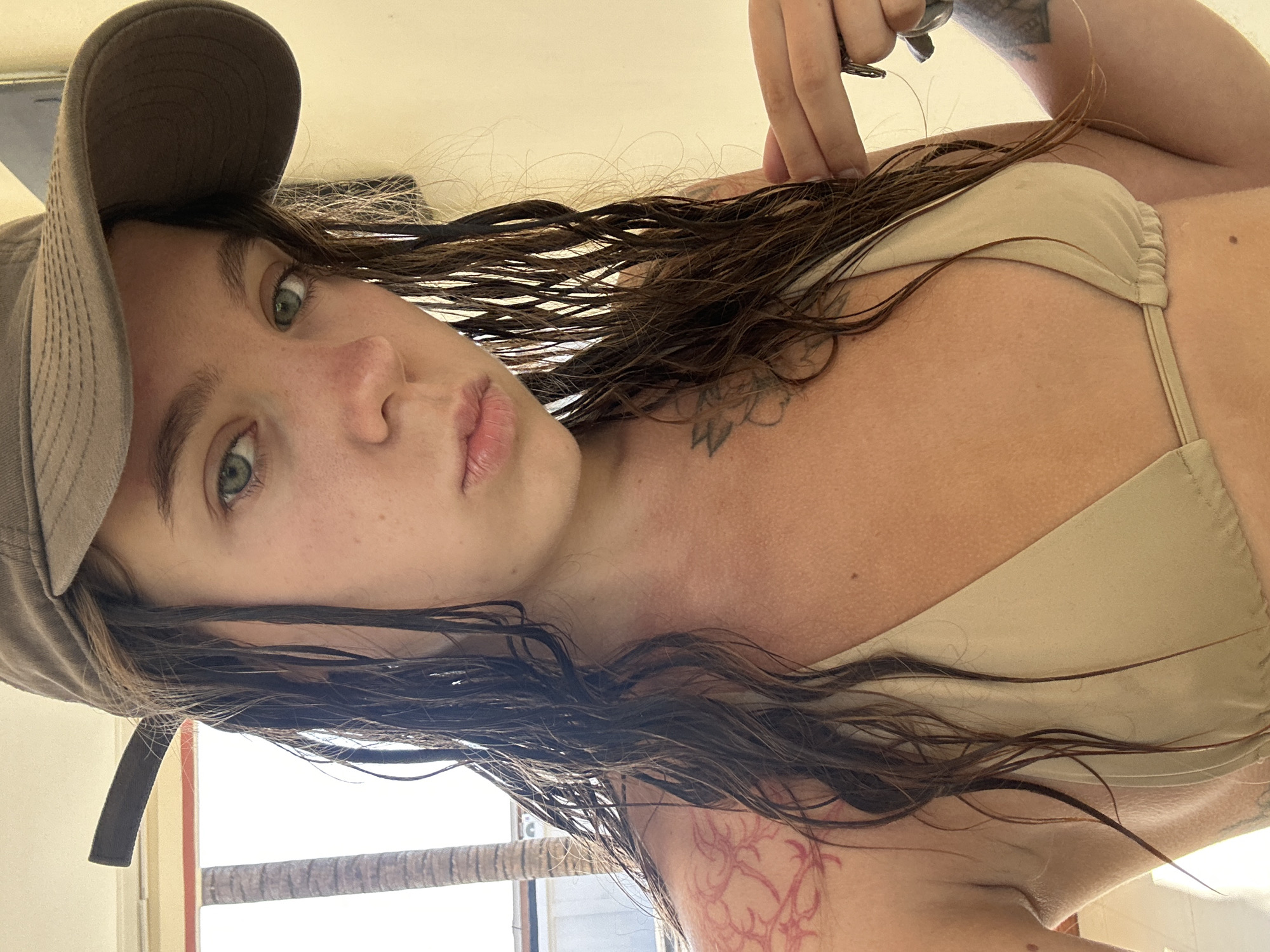 A young woman with wet brown hair, wearing a grey baseball cap and a beige bikini top, is pictured from the chest up. She has light skin, green eyes, and a smattering of freckles on her face. A reddish tattoo is visible on her left shoulder, and other tattoos are present on her torso and arm. Her right hand is near her face, holding onto some keys. The background is out of focus but appears to be an indoor setting with a window, a palm tree visible outside, and a light-colored wall. The lighting is bright and natural.
