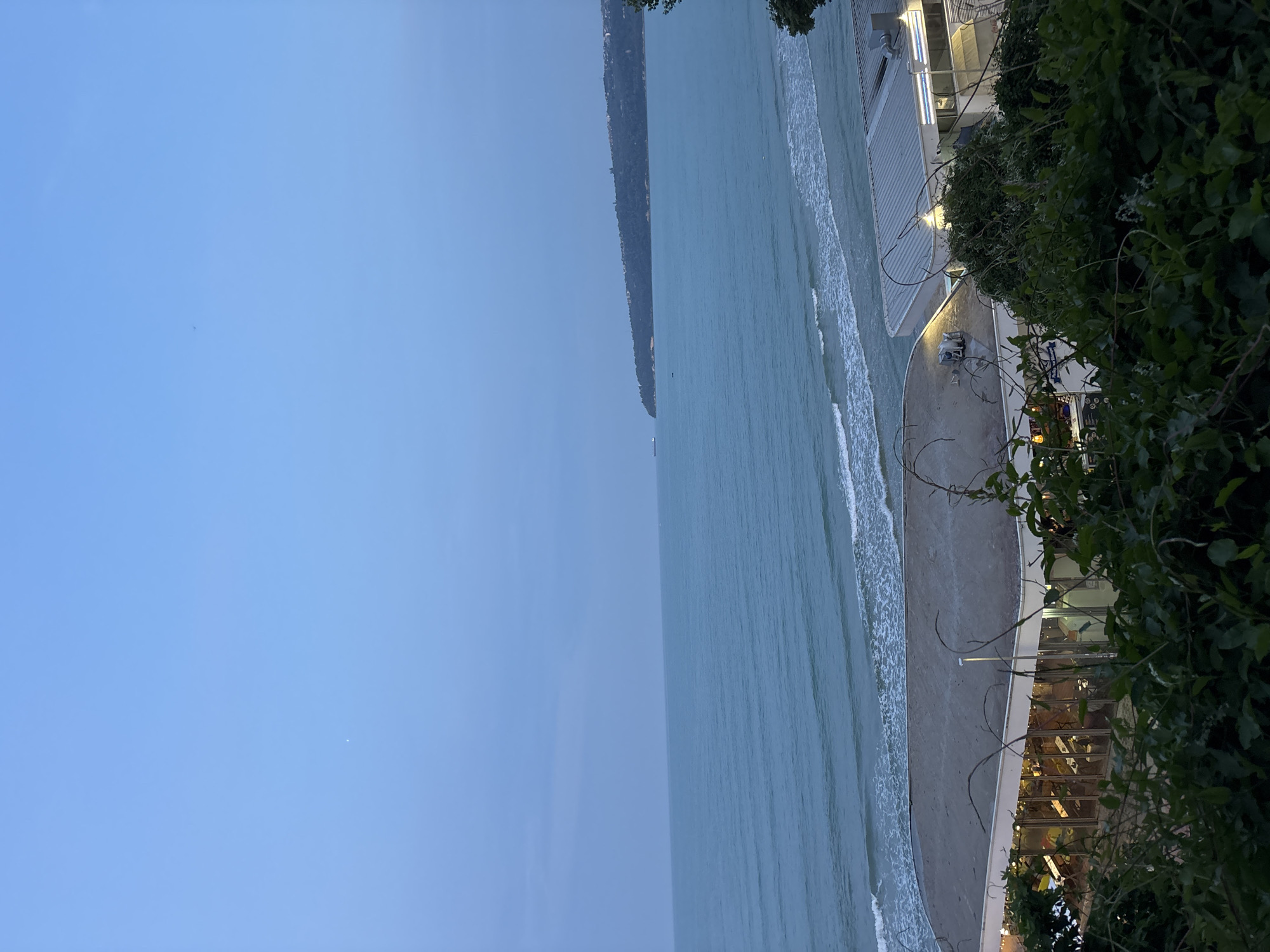 The image presents a serene coastal scene, likely along the Black Sea in Varna, Bulgaria, captured during the late afternoon or early evening. The sky is a clear, light blue, gradually fading into the dusk.
The central feature is the calm, light blue-green sea with gentle waves breaking onto a narrow strip of sandy beach. Across the water, a long, dark, forested cliff or landmass stretches along the horizon, adding depth to the panorama.
On the right side of the image, modern, white-facade buildings with large glass windows stand directly adjacent to the beach. These structures appear to be restaurants or cafes, with some outdoor seating and illuminated interiors. Artificial lights are visible, indicating the approach of evening. Lush, dark green foliage, including bushes and vines, frames the foreground on the right, partially obscuring the buildings and the beach.
No prominent human activity is taking place, suggesting a peaceful and quiet atmosphere. A small, dark object, possibly a distant boat or person, is faintly visible in the water. One of the buildings has a blue sign with the word "CLUB" partially visible. The overall mood is tranquil, characterized by clear weather and calm waters.