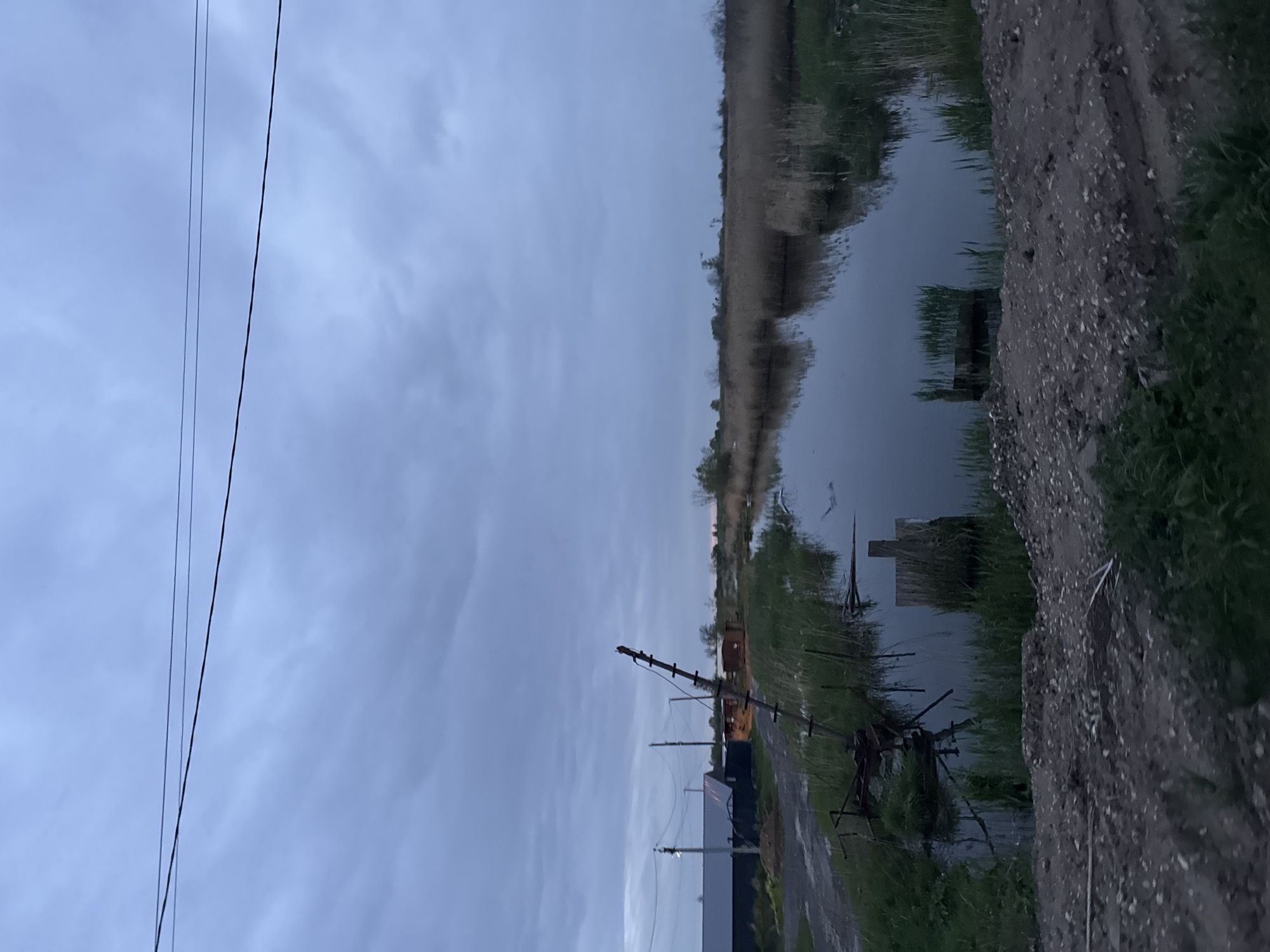The image shows a canal-side landscape in an unspecified urban location. The sky is overcast and the time appears to be dusk or dawn. A body of water, possibly a canal or river, runs horizontally across the top third of the frame, its surface reflecting the sky. The opposite bank is lined with vegetation, possibly reeds or tall grasses. The lower portion of the image shows the canal bank, with a gravel or dirt path running along it. There are various pieces of metal debris or rusting structures near the water's edge. In the distance, utility poles and wires are visible against the sky, suggesting an urban or industrial backdrop. The lighting is dim, creating a somber atmosphere.