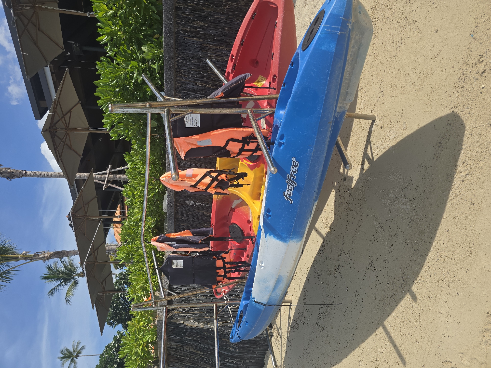 In Baan Chaweng Noi, Thailand, a sunny beach scene features two kayaks on a sandy shore. A blue and white kayak is in the foreground, supported by wooden legs. Behind it, a yellow kayak is partially visible, and a red kayak is further back. Several orange life vests and a black life vest are hanging from a metal rack positioned above the kayaks. In the background, lush green foliage lines a stone wall, with large umbrellas providing shade over an outdoor seating area of a building. Palm trees are visible against a clear blue sky with scattered clouds.